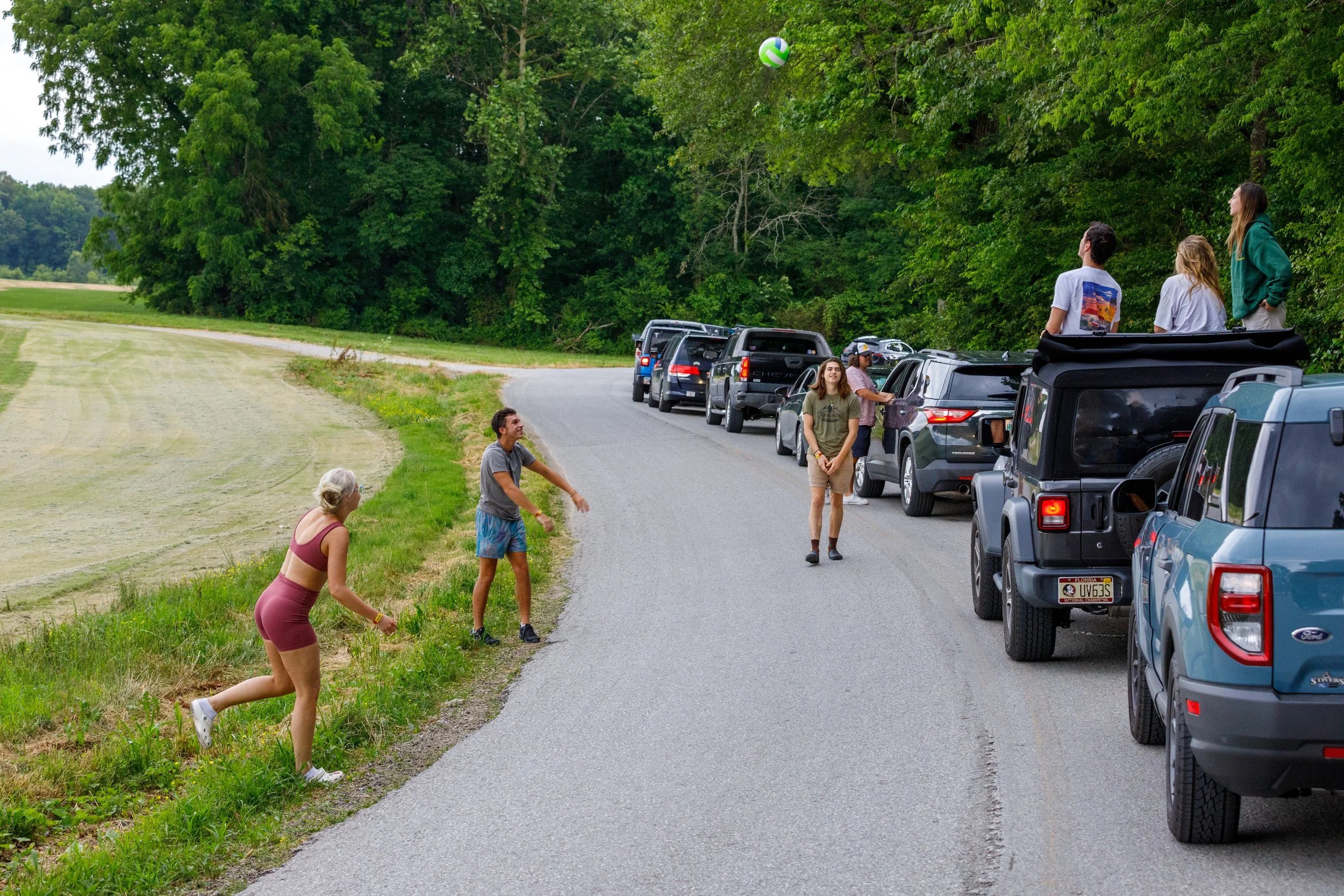 People playing volleyball on a country road next to a line of parked cars with trees in the background.