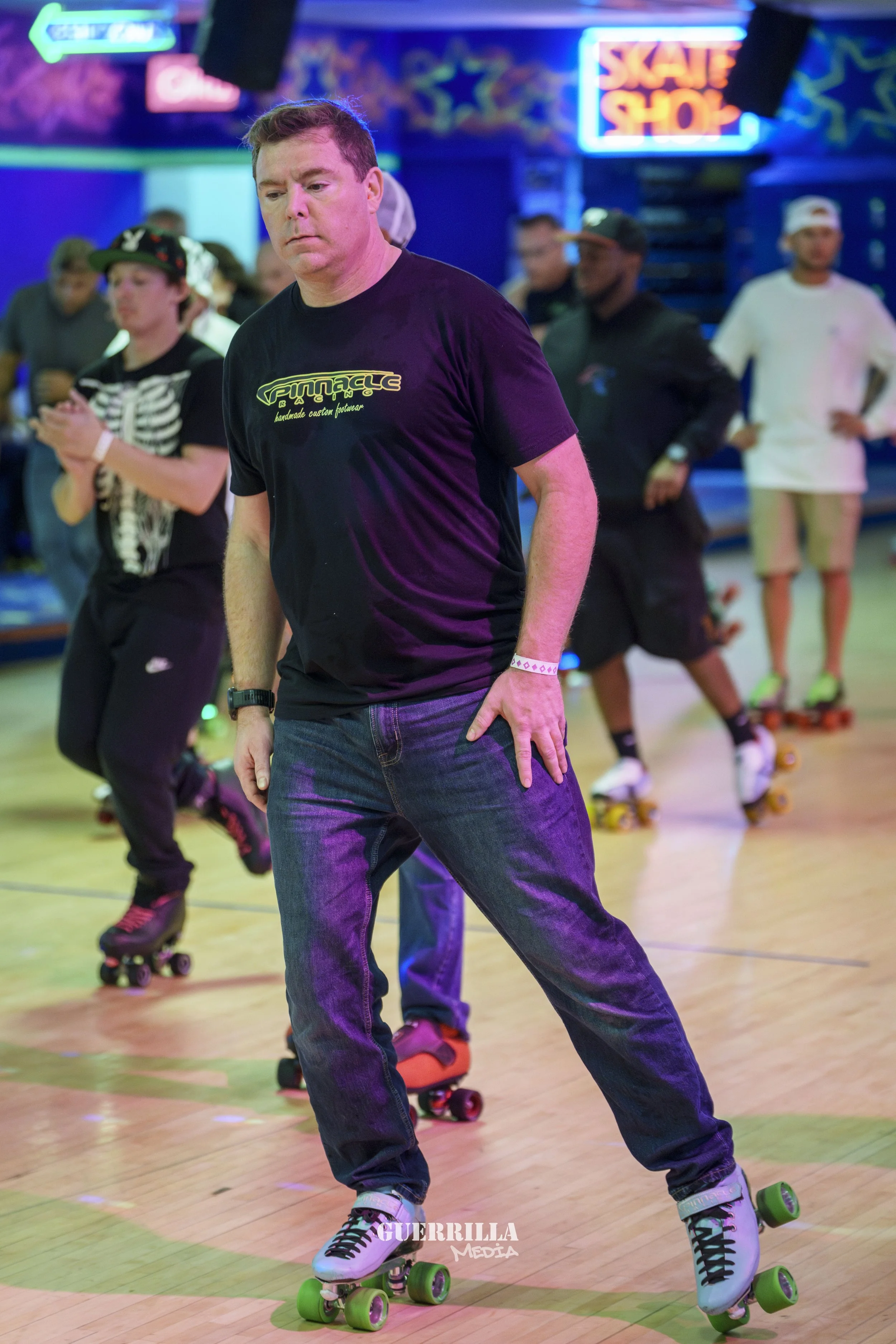 A man roller skating in an indoor roller rink with neon signs and other skaters in the background.