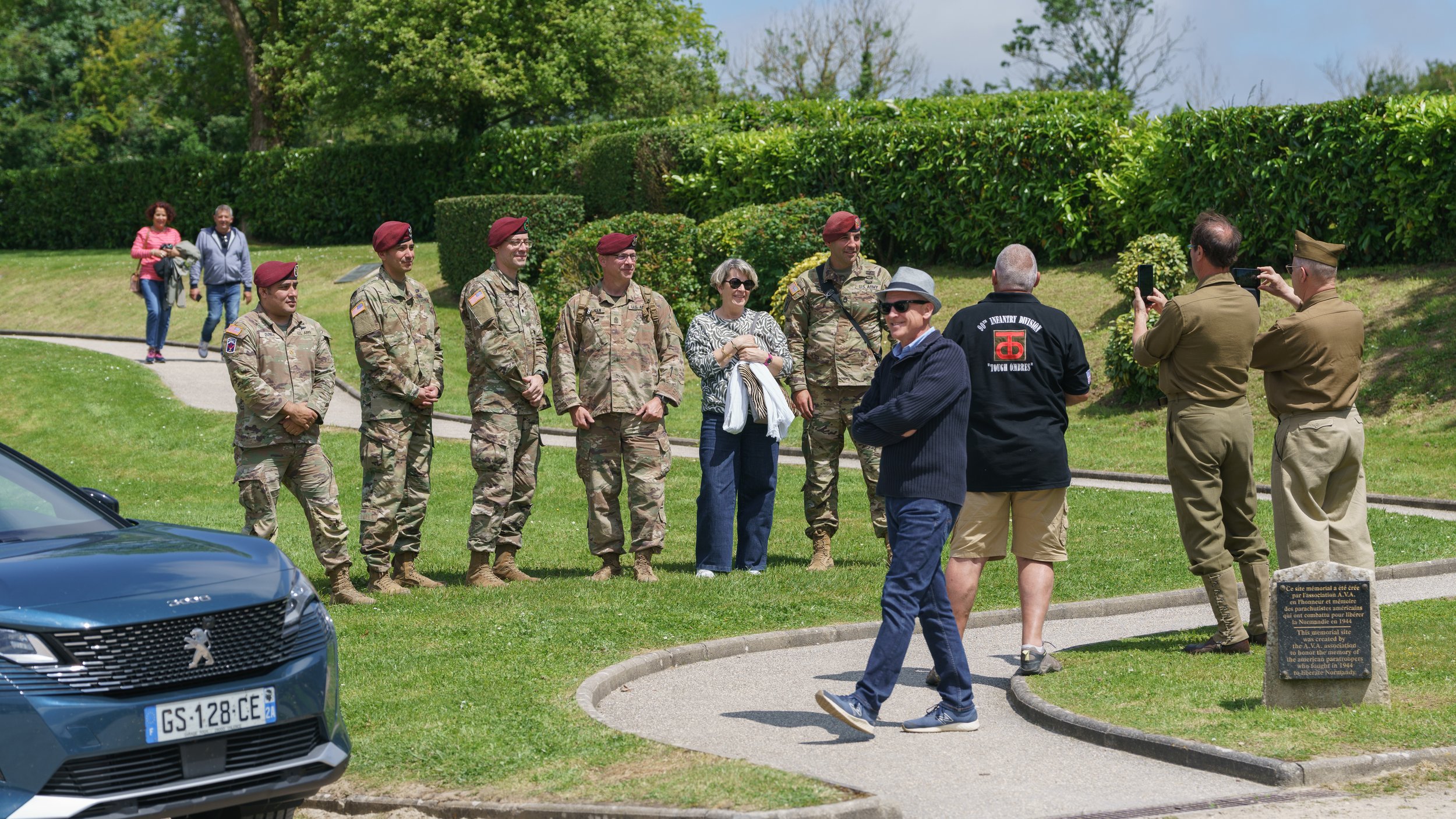 Group of military personnel and civilians standing outdoors in front of green bushes, with some taking photos and others chatting, during a visit or tour.