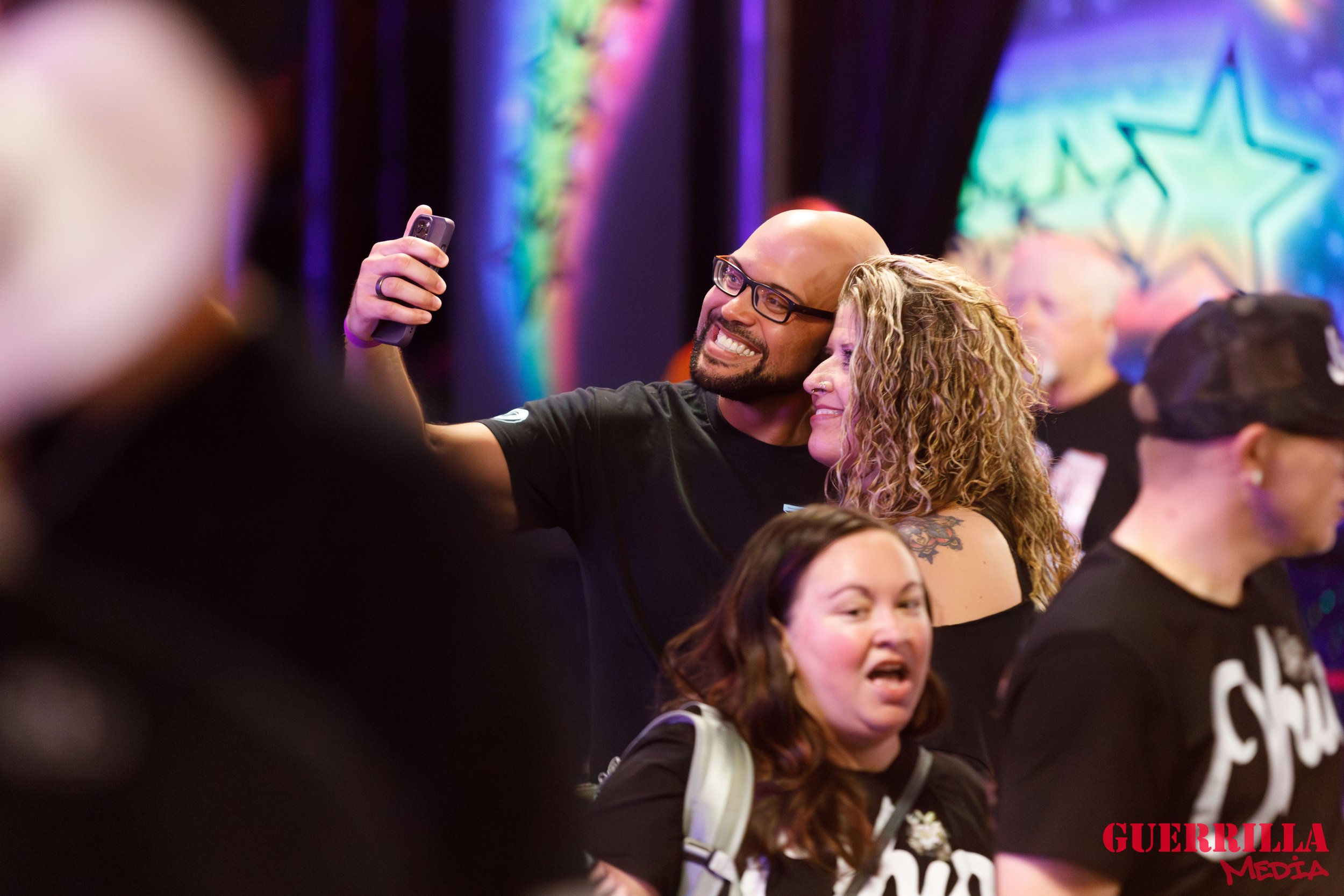 Group of people at an event, with a man in glasses taking a selfie with a woman who has curly hair, both smiling. A woman with brown hair and a man in a black baseball cap are also in the crowd. Colorful neon lighting in the background.