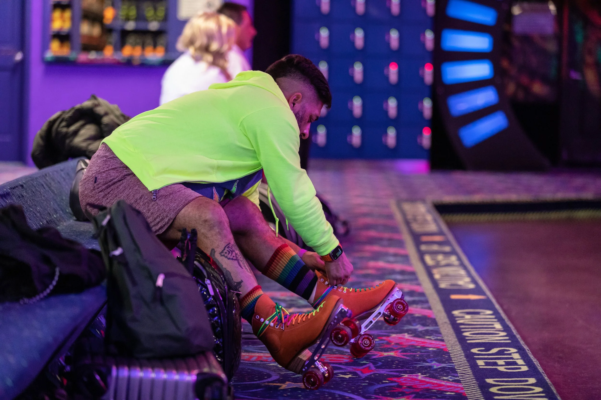 Person adjusting orange roller skates with rainbow-colored laces, dressed in a neon green jacket and rainbow socks, sitting on a bench in a colorful indoor roller skating rink.