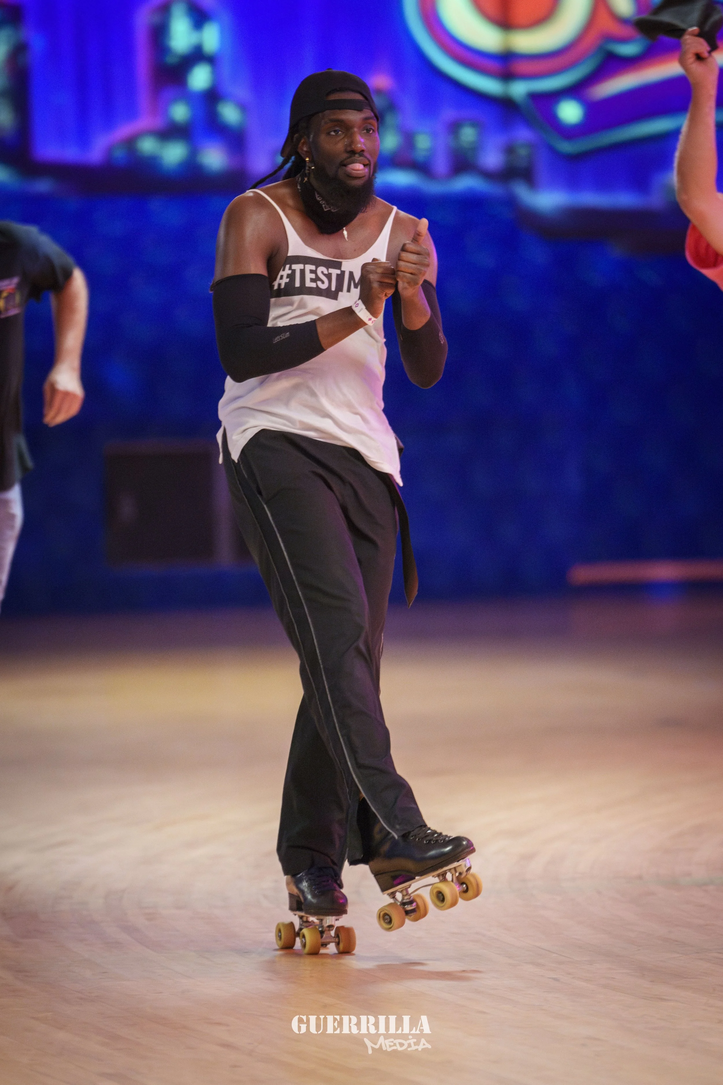 Person skating indoors at an event with a colorful mural background, wearing athletic clothing, a cap, and arm sleeves, during a roller skating or dance activity.