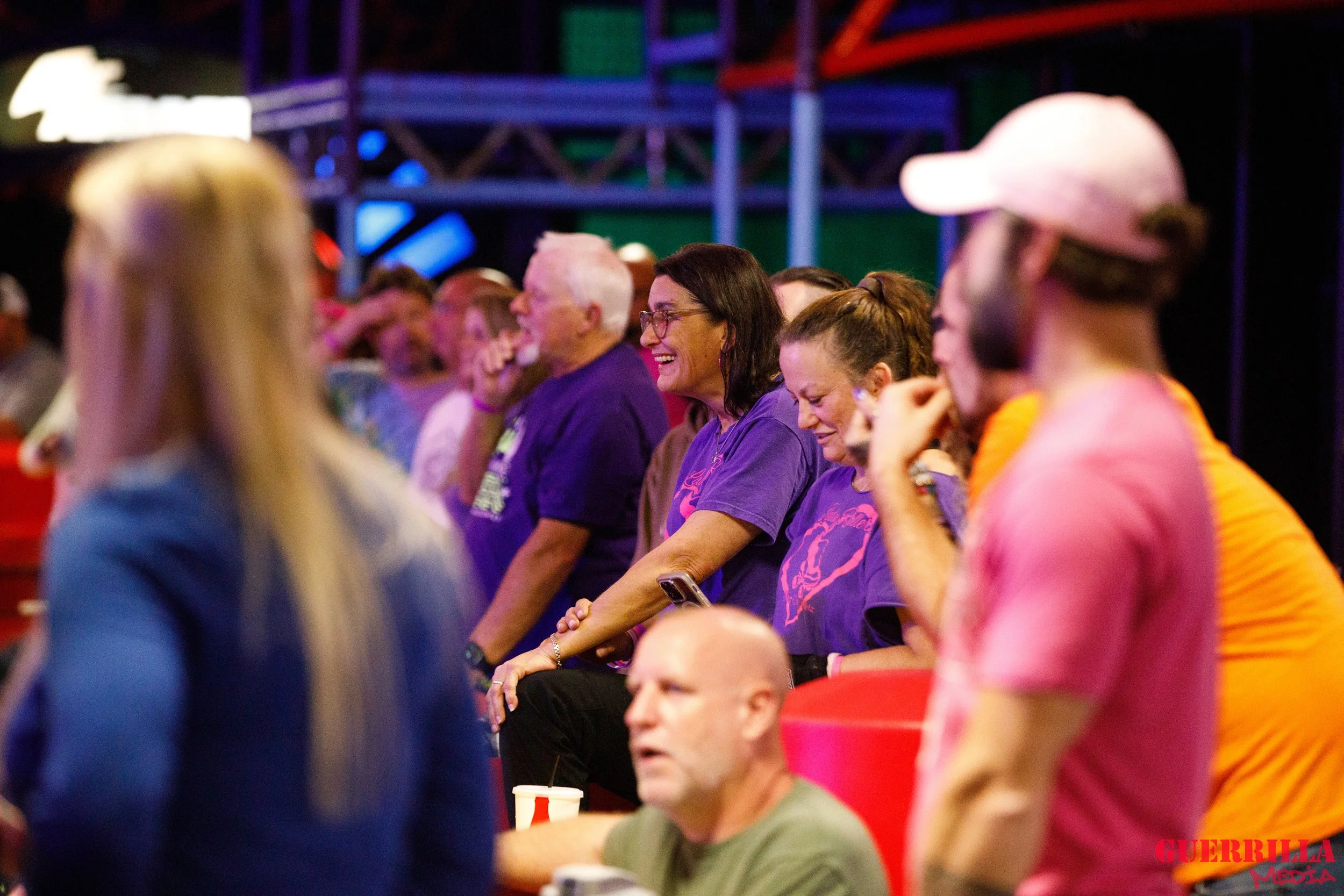People sitting in a row at an indoor event, some laughing or looking amused, with colorful lighting and a steel framework in the background.