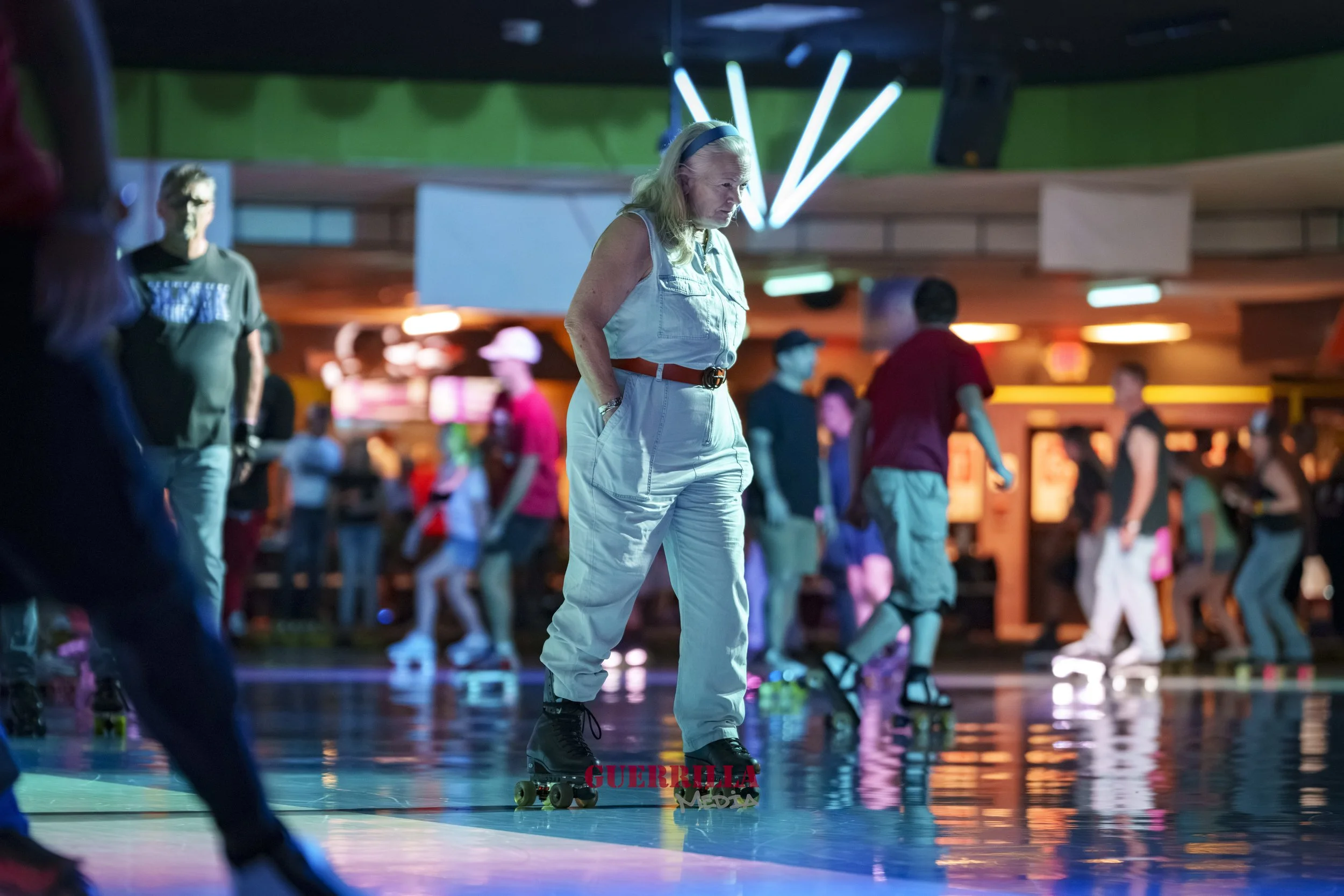 An elderly woman roller skating at an indoor skating rink, surrounded by other skaters in the background.