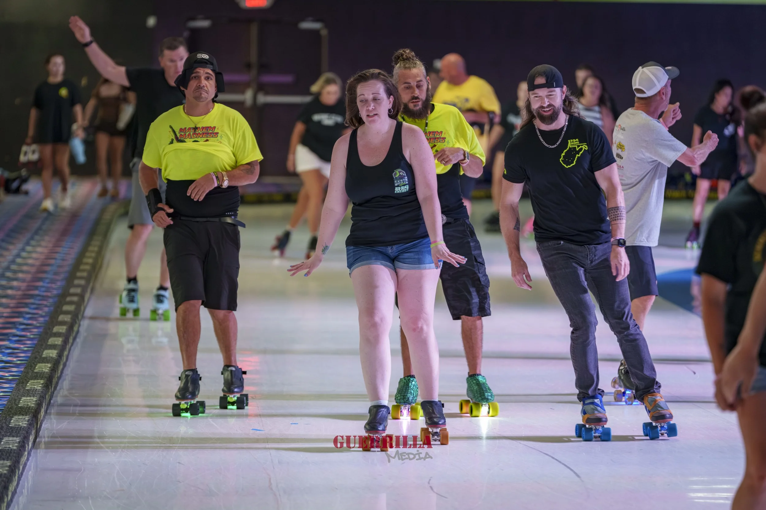 Group of people roller skating indoors, some wearing bright yellow shirts, others in casual clothing, with a woman in black shorts and tank top at the front, some smiling and others with focused expressions.
