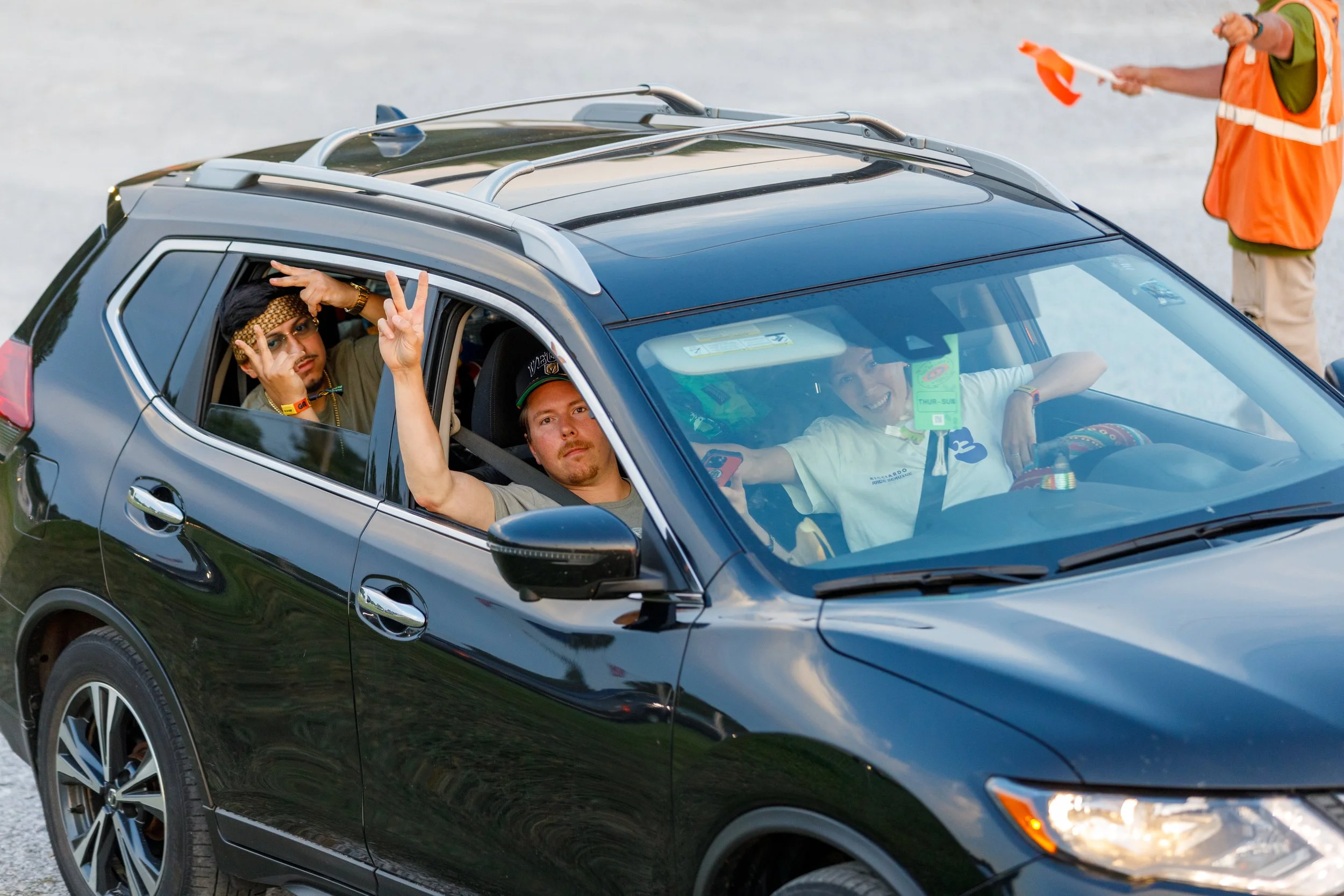 People sitting inside a black SUV on a racetrack, with one person in the driver's seat and two passengers in the back. The passengers are making peace signs and posing for the camera. There is a person outside the vehicle holding a flag, and a person