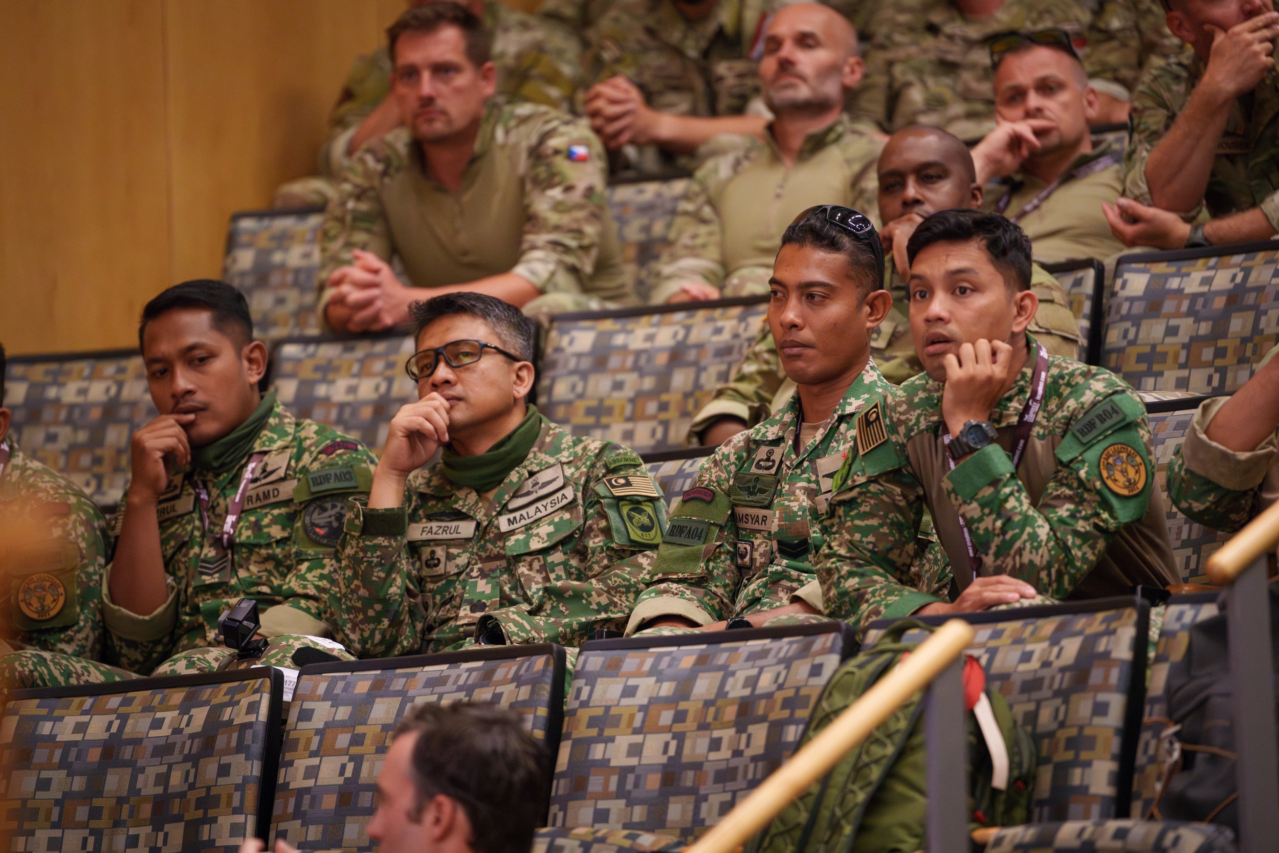 Group of soldiers in camouflage uniforms sitting in an auditorium during a presentation or training session.