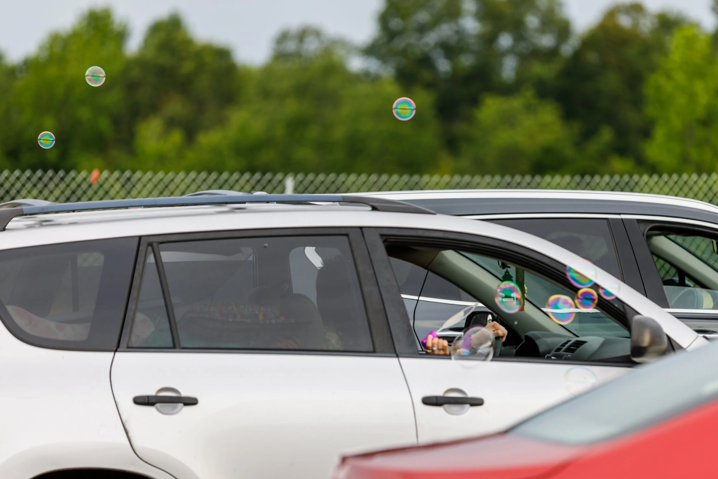 A white SUV with a person inside holding a mobile phone, parked outdoors near a fence. There are soap bubbles floating in the air around the vehicle and in the background, which features green trees.