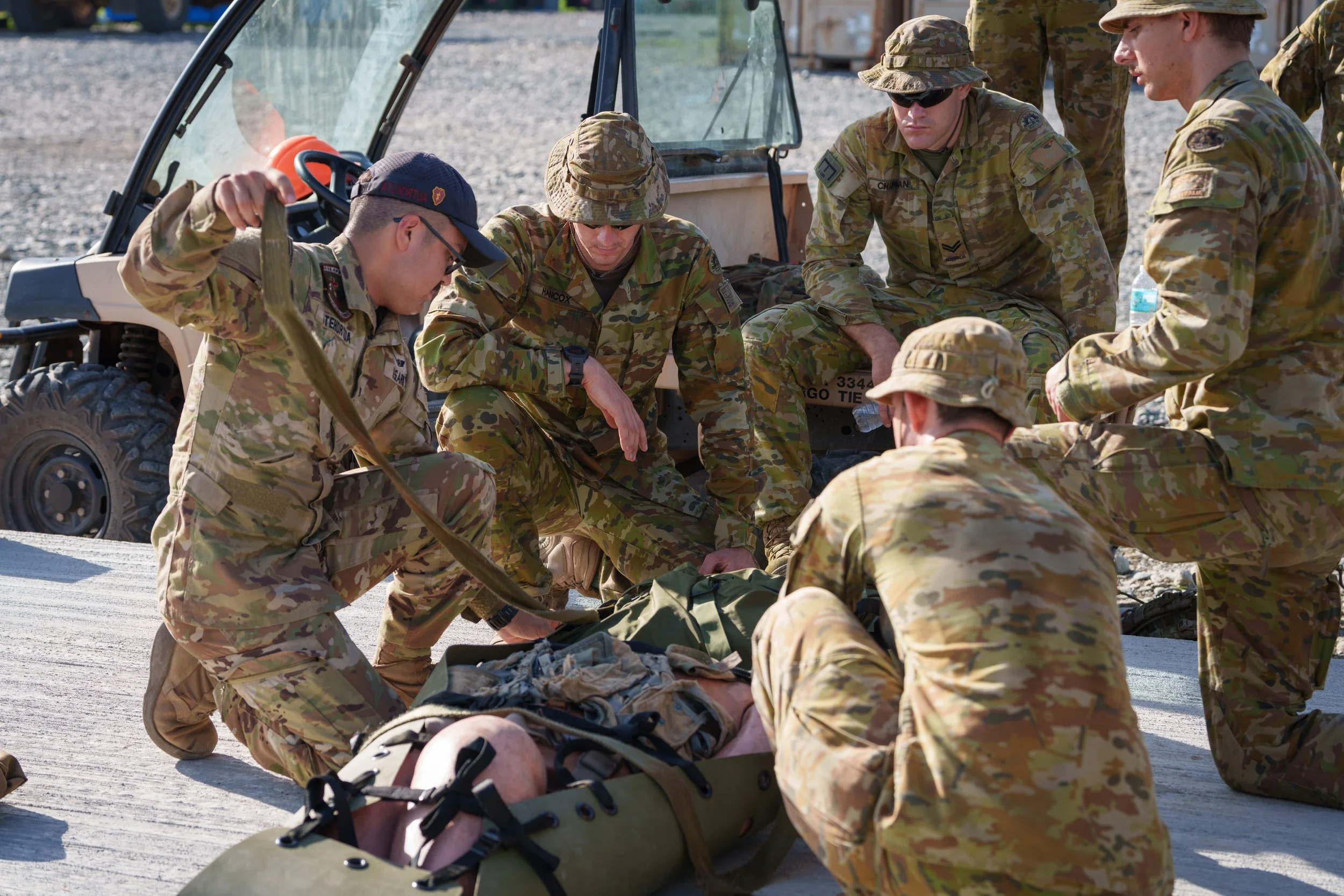 Military personnel performing first aid or medical training on a person lying on the ground, with a military vehicle in the background.