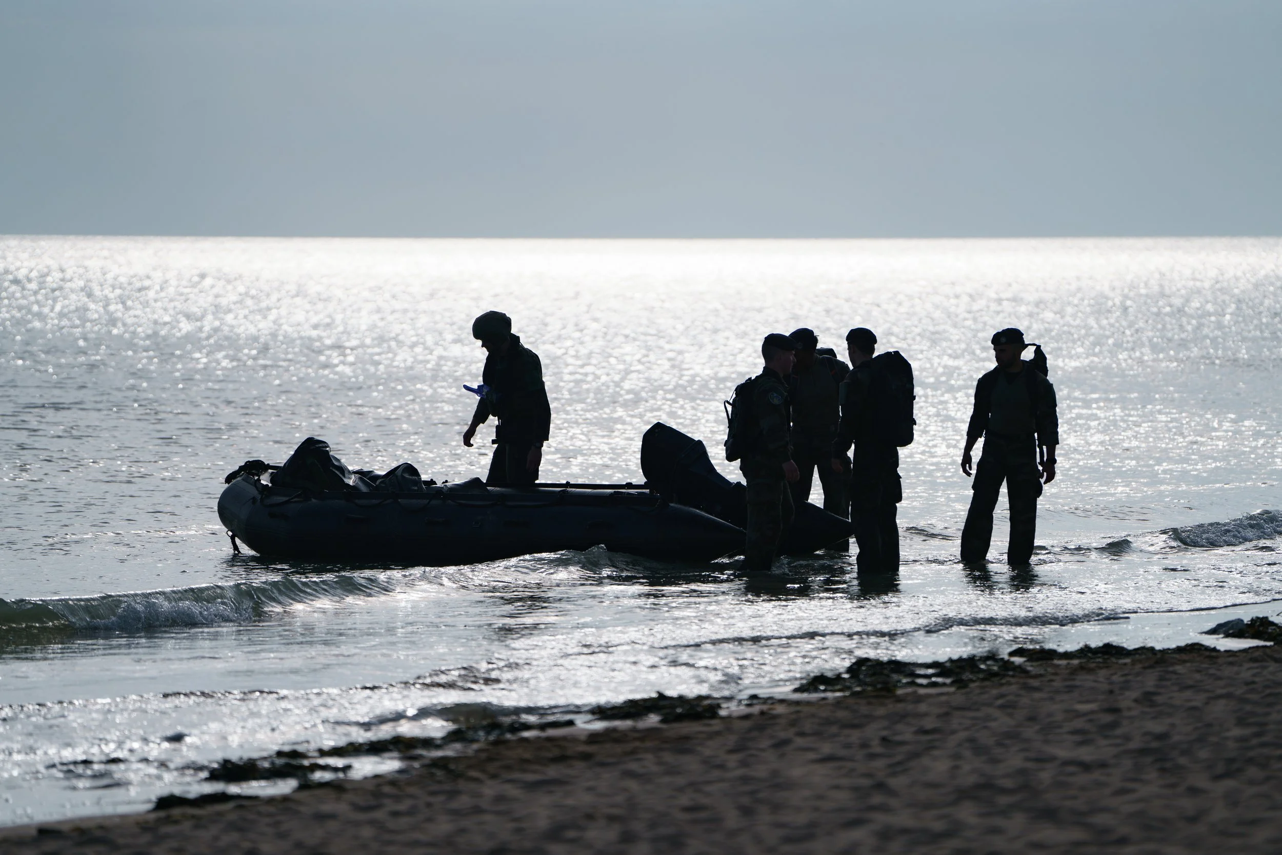 Five soldiers in uniform on a beach, managing a small inflatable boat near the water.