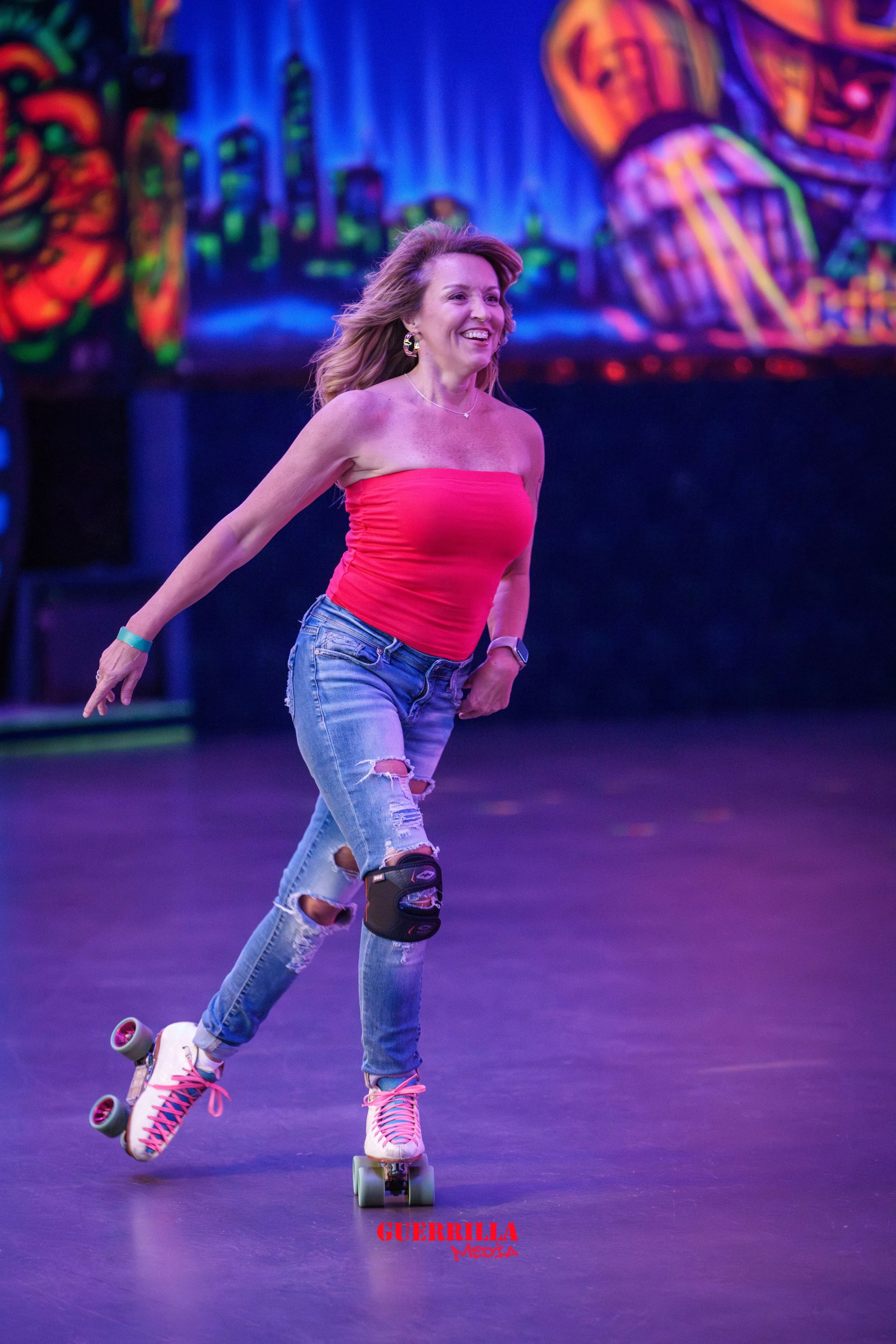 A woman roller skating indoors with a spoke smile, wearing a red strapless top, ripped jeans, and pink laced roller skates. Brightly colored art decorates the background wall.