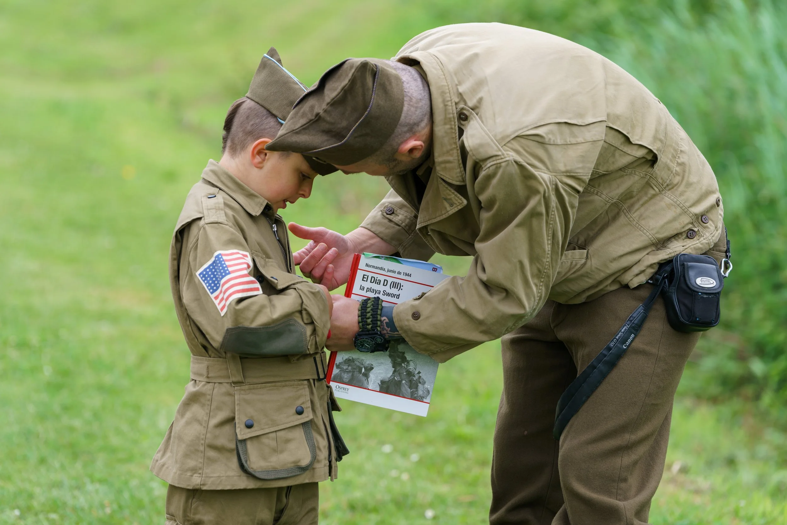 A man and a young boy dressed in military-style uniforms, with the boy wearing a patch of the American flag on his sleeve, are outdoors on a grassy area. The man is leaning over, showing the boy a book titled "El Día D (III): la playa Sword".