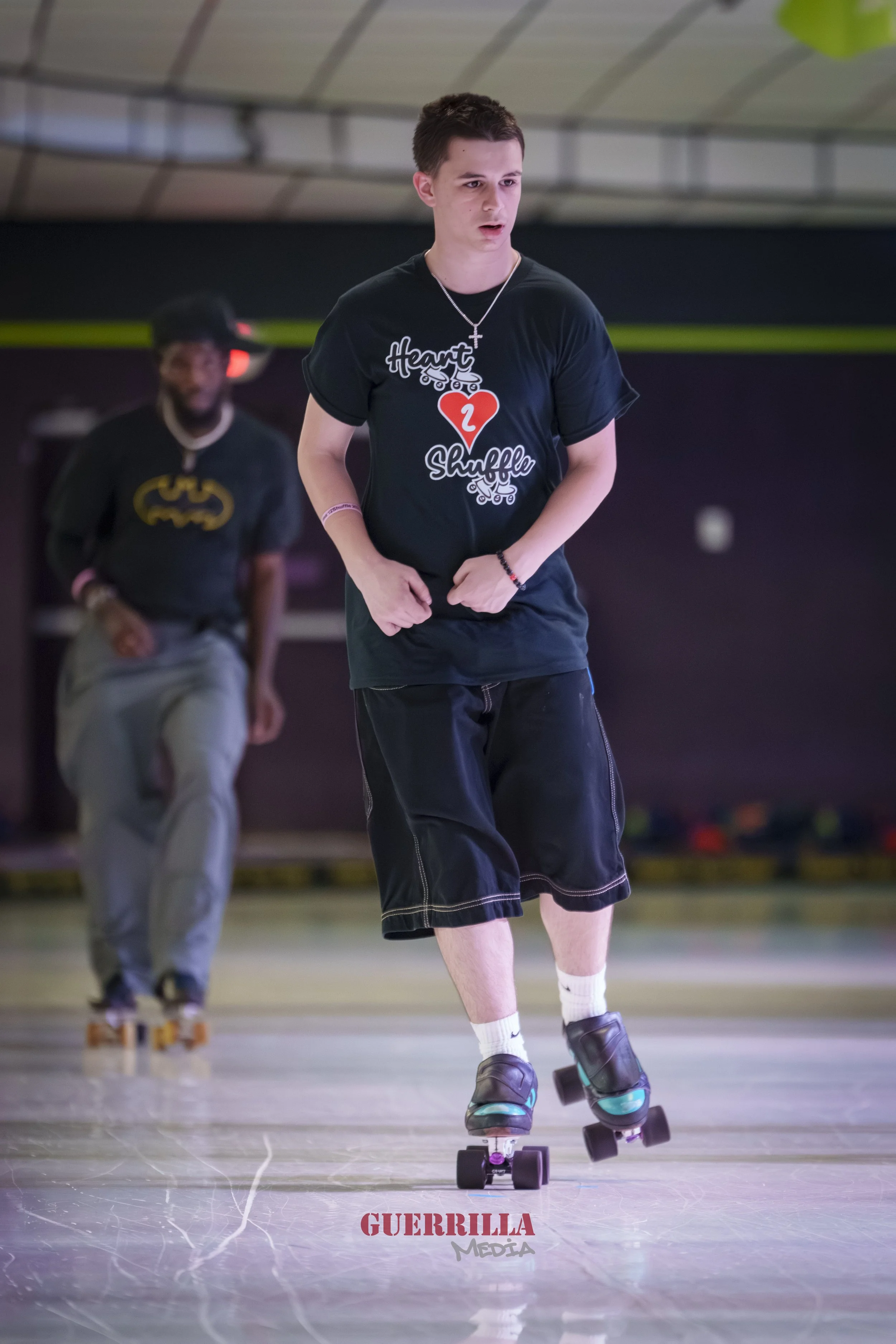 Young man roller skating indoors, wearing a black t-shirt with the words 'Heart Shuffle' and a red heart, black shorts, white socks, and roller skates. Another person is in the background, also roller skating.