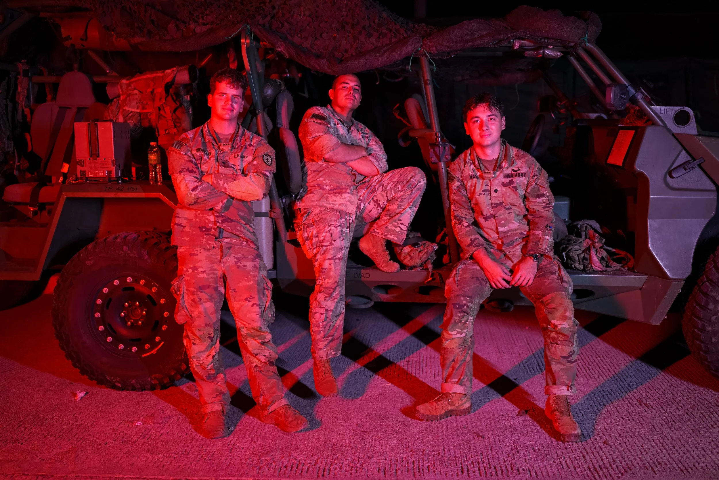 Three soldiers in camouflage uniforms standing and sitting in front of a military vehicle, illuminated with red light.