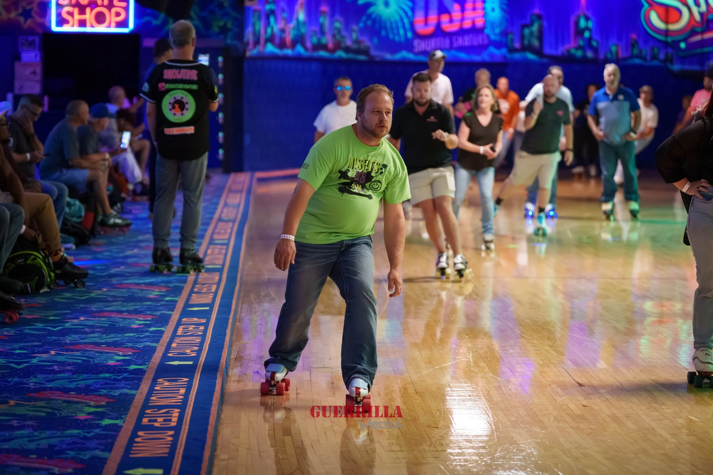 People roller skating inside an indoor roller rink with colorful lights and neon signs, with some spectators sitting and standing along the sides.