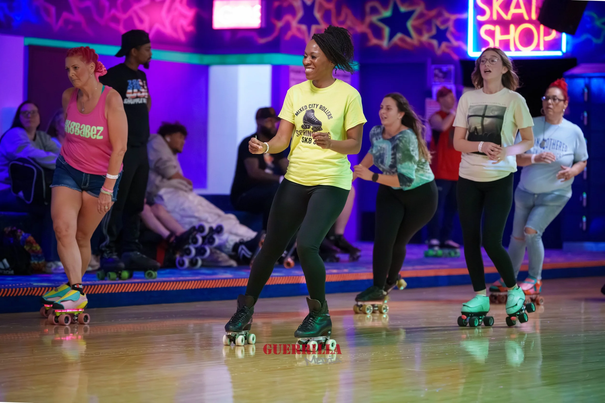 A group of women roller skating inside a brightly decorated skating rink with neon signs and colorful lights.