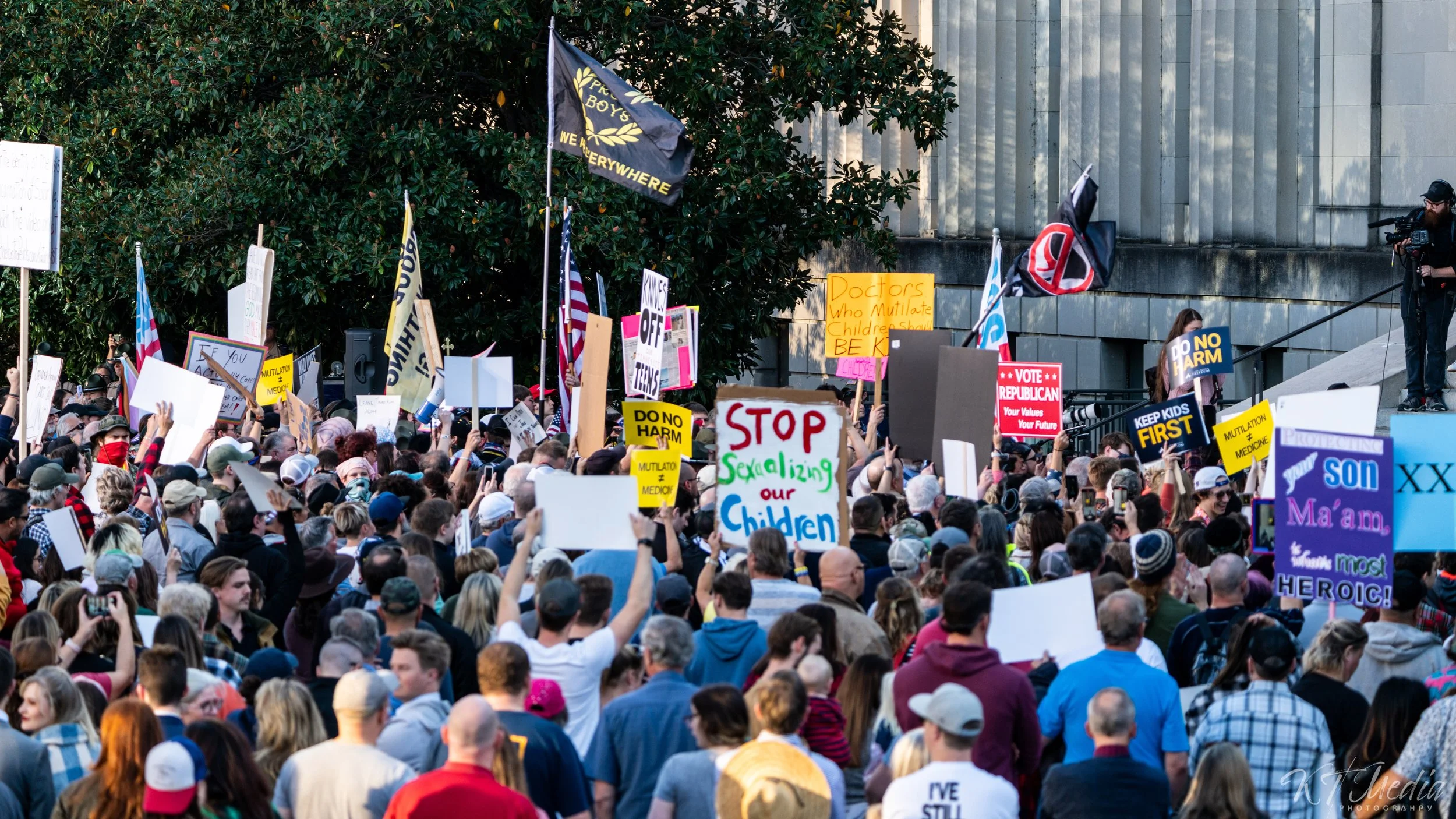 A large crowd of people gathered at a protest, holding various signs and banners. Some signs advocate against harm to children and emphasize prioritizing kids' needs. Flags and a cameraman are also visible.