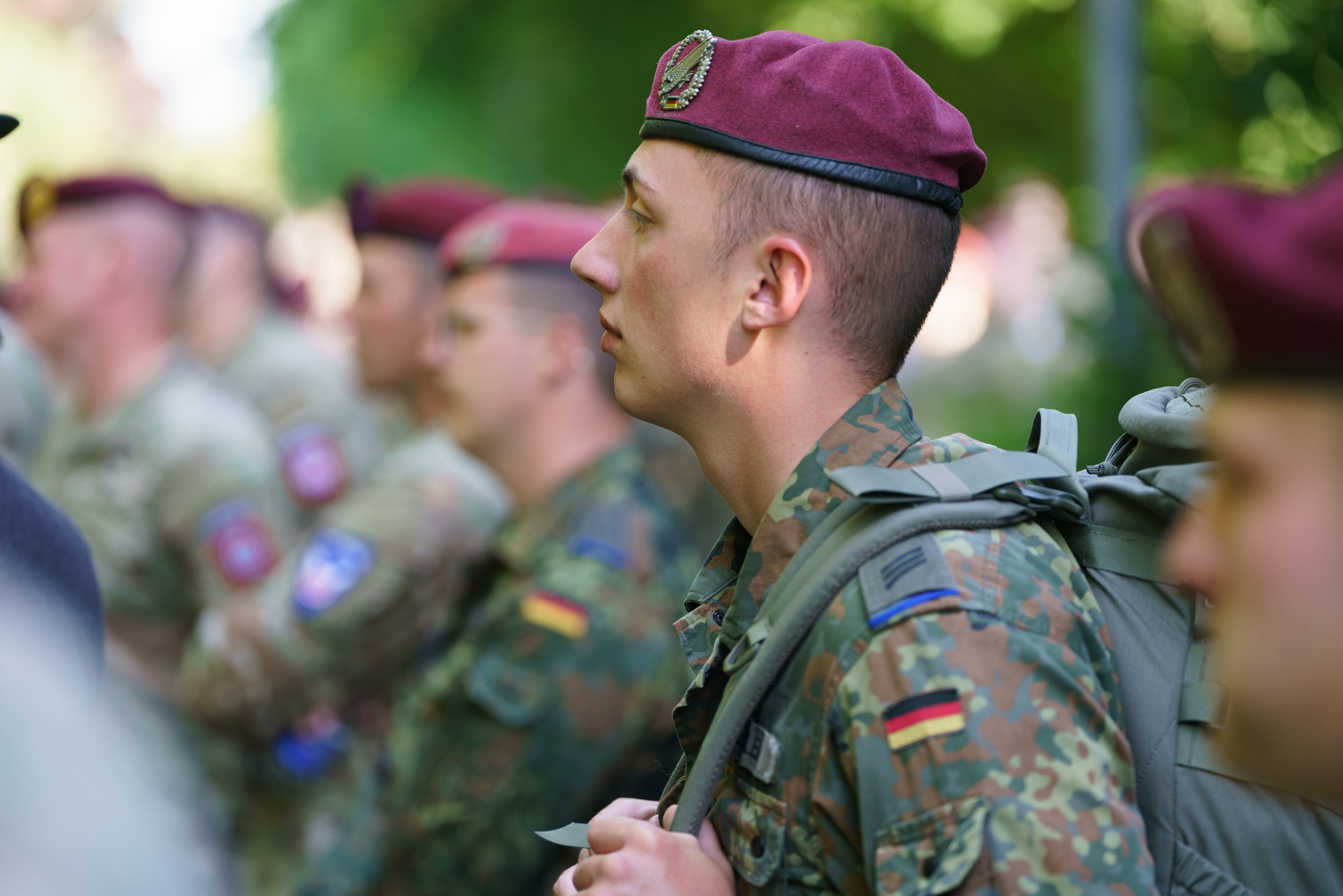 A group of soldiers standing in a line outdoors, wearing camouflage uniforms and maroon berets, with some patches and flags on their sleeves.