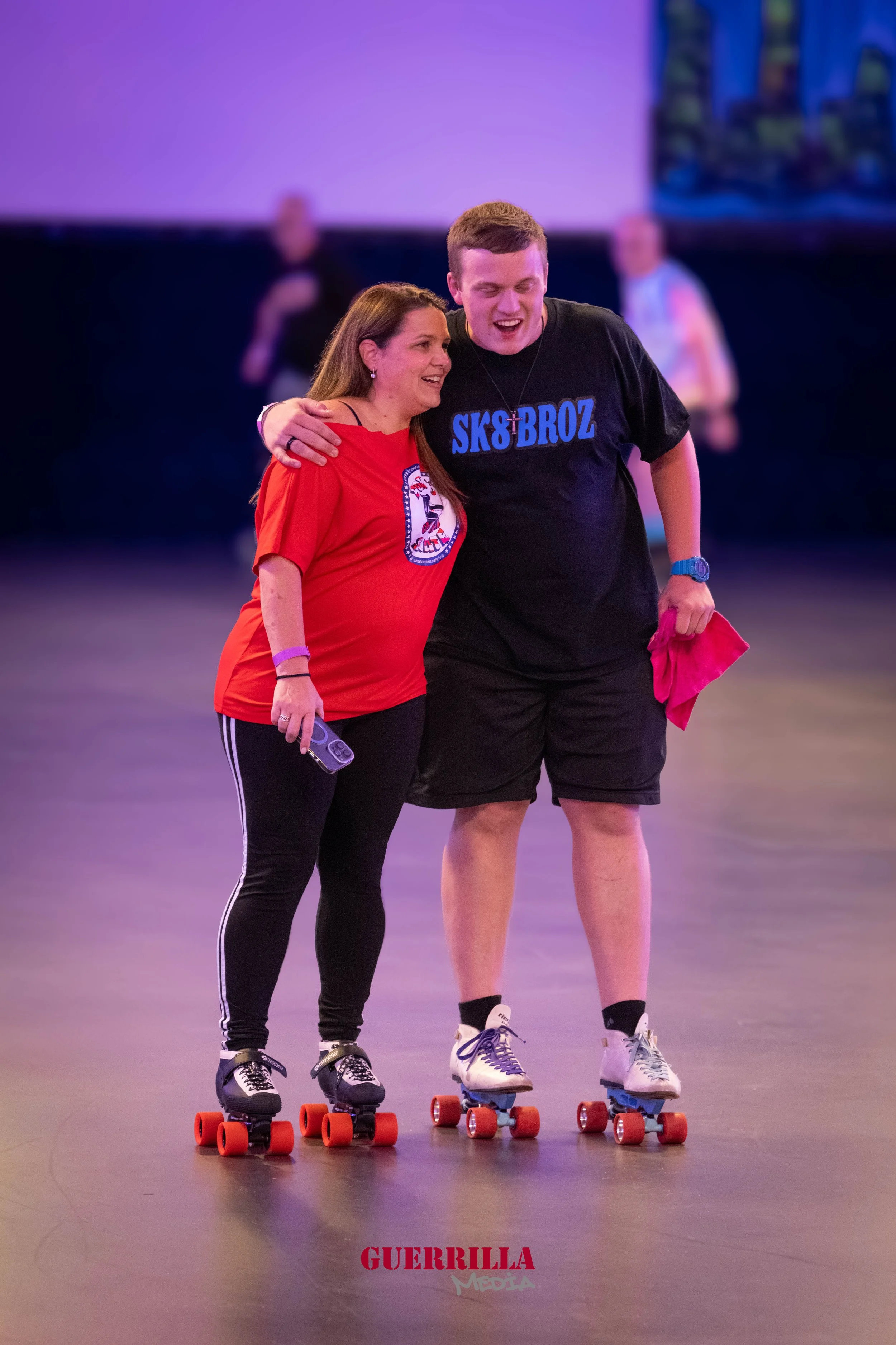 Two people roller skating together, smiling, with the woman wearing a red shirt and the man wearing a black shirt with blue text, holding a pink cloth, in a roller rink with purple and blue lighting.