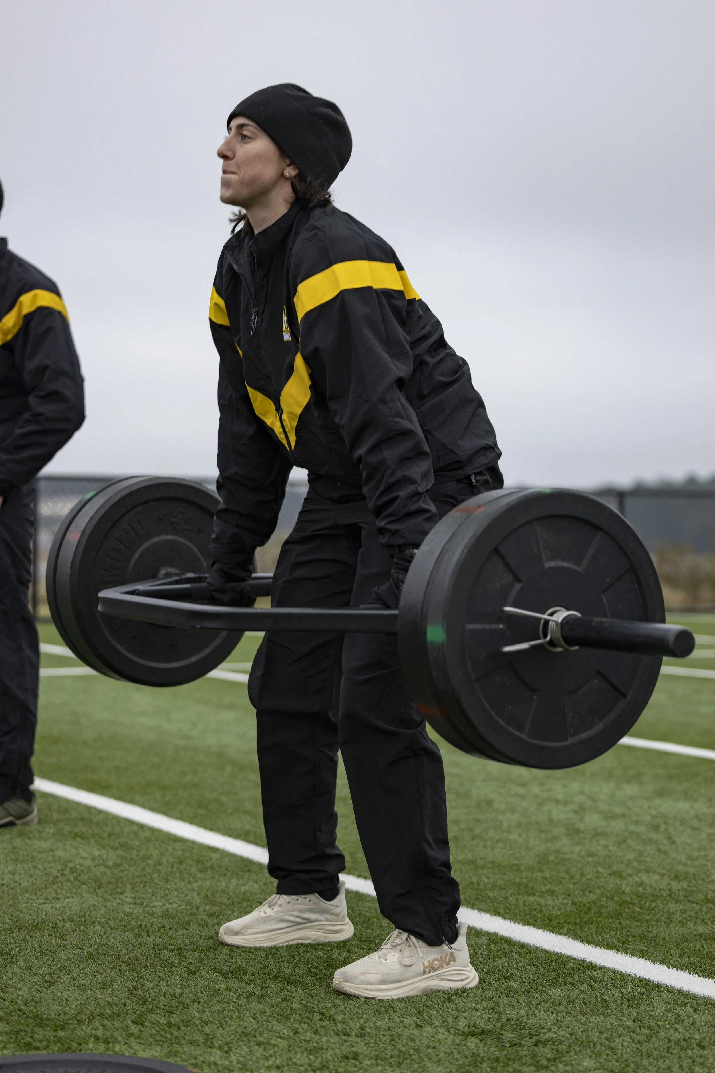 A woman in black athletic gear with a yellow stripe, wearing a black beanie, is lifting a barbell with large black weights outdoors on a sports field.