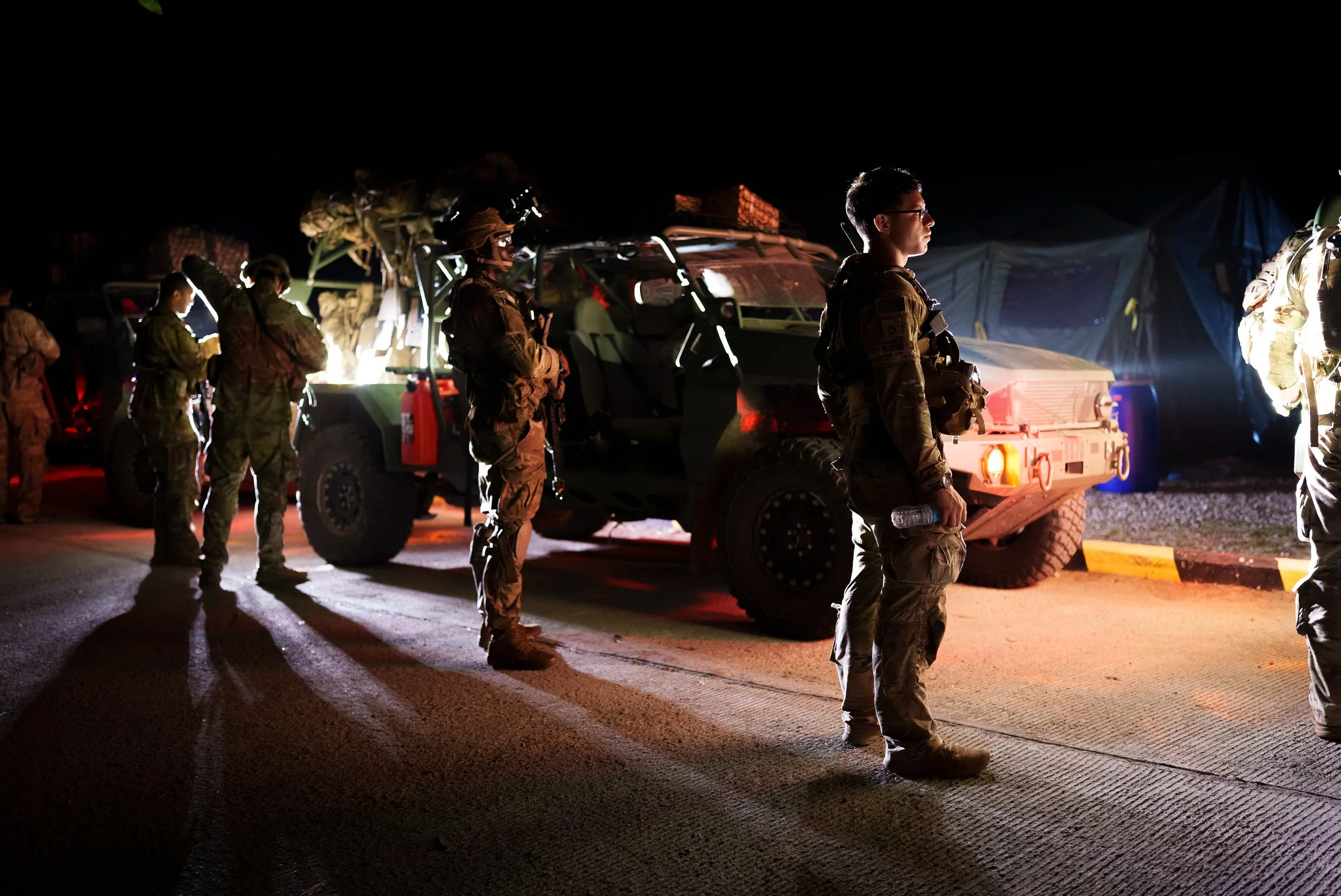 Military personnel standing in line at night next to military vehicles under artificial lights.