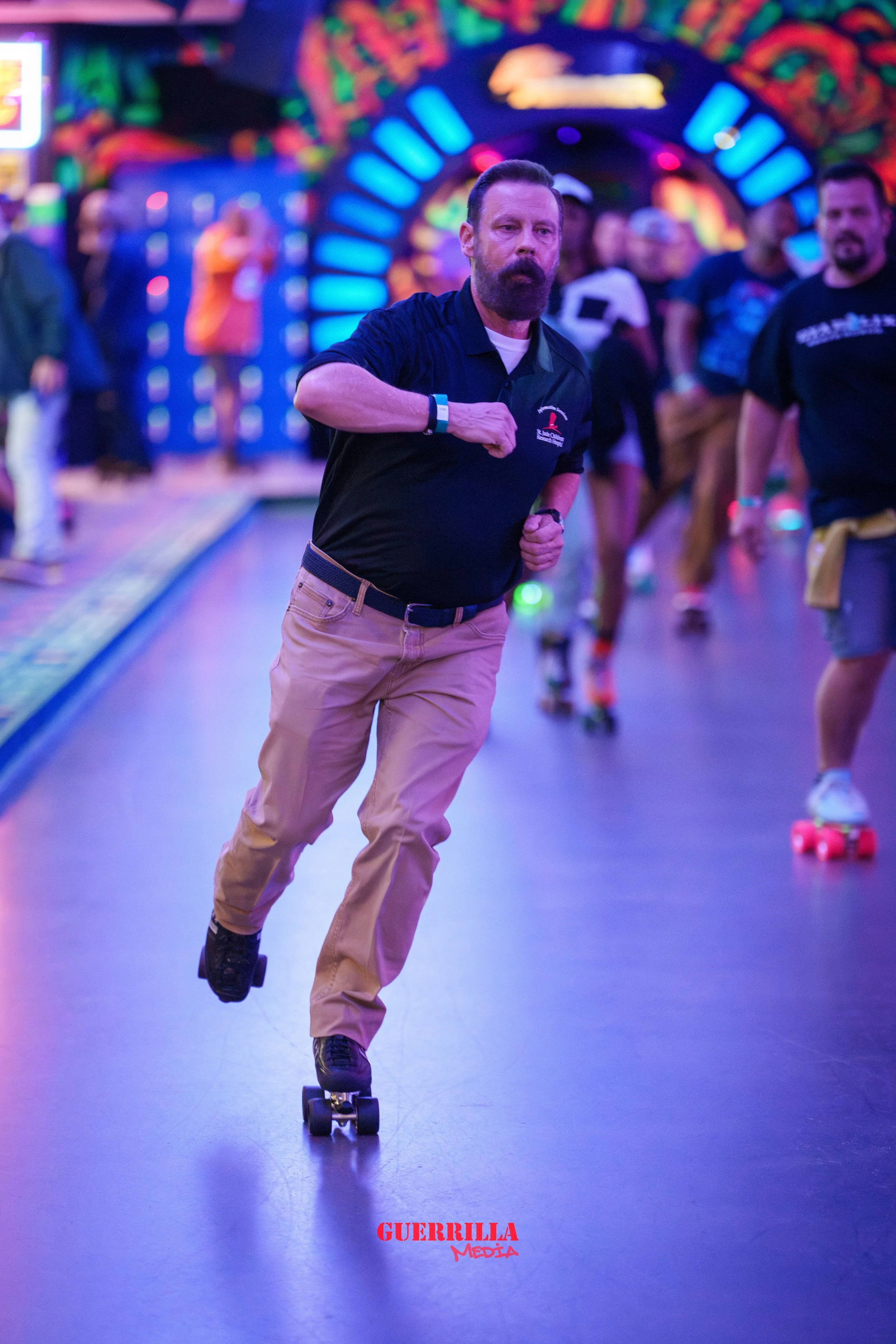A man roller skating in an indoor amusement park with bright, colorful neon lights and other skaters in the background.