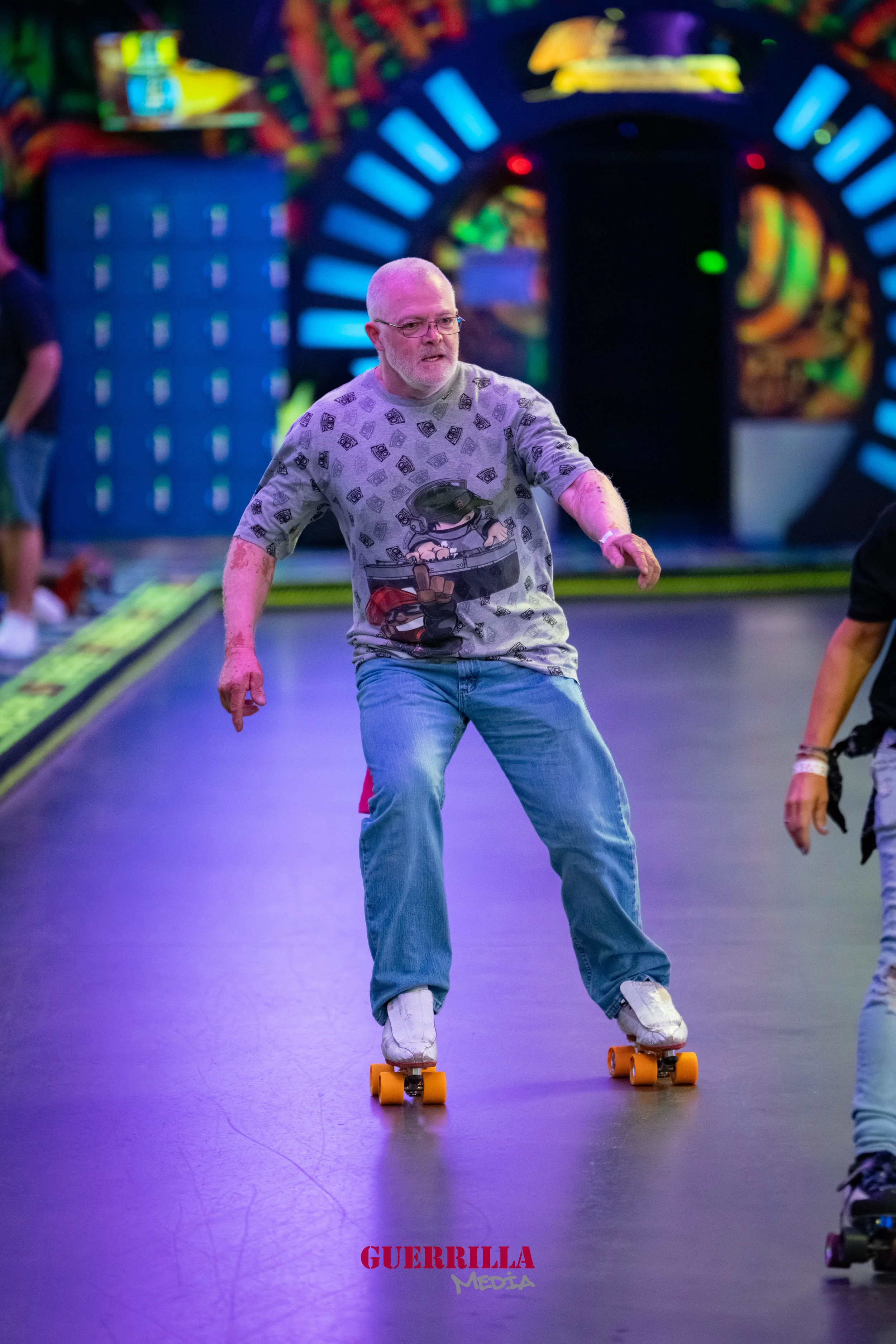 A man with gray hair and glasses is roller skating in an indoor arcade or skating rink, wearing a casual t-shirt with cartoon graphics and jeans, surrounded by colorful arcade games and bright lights.