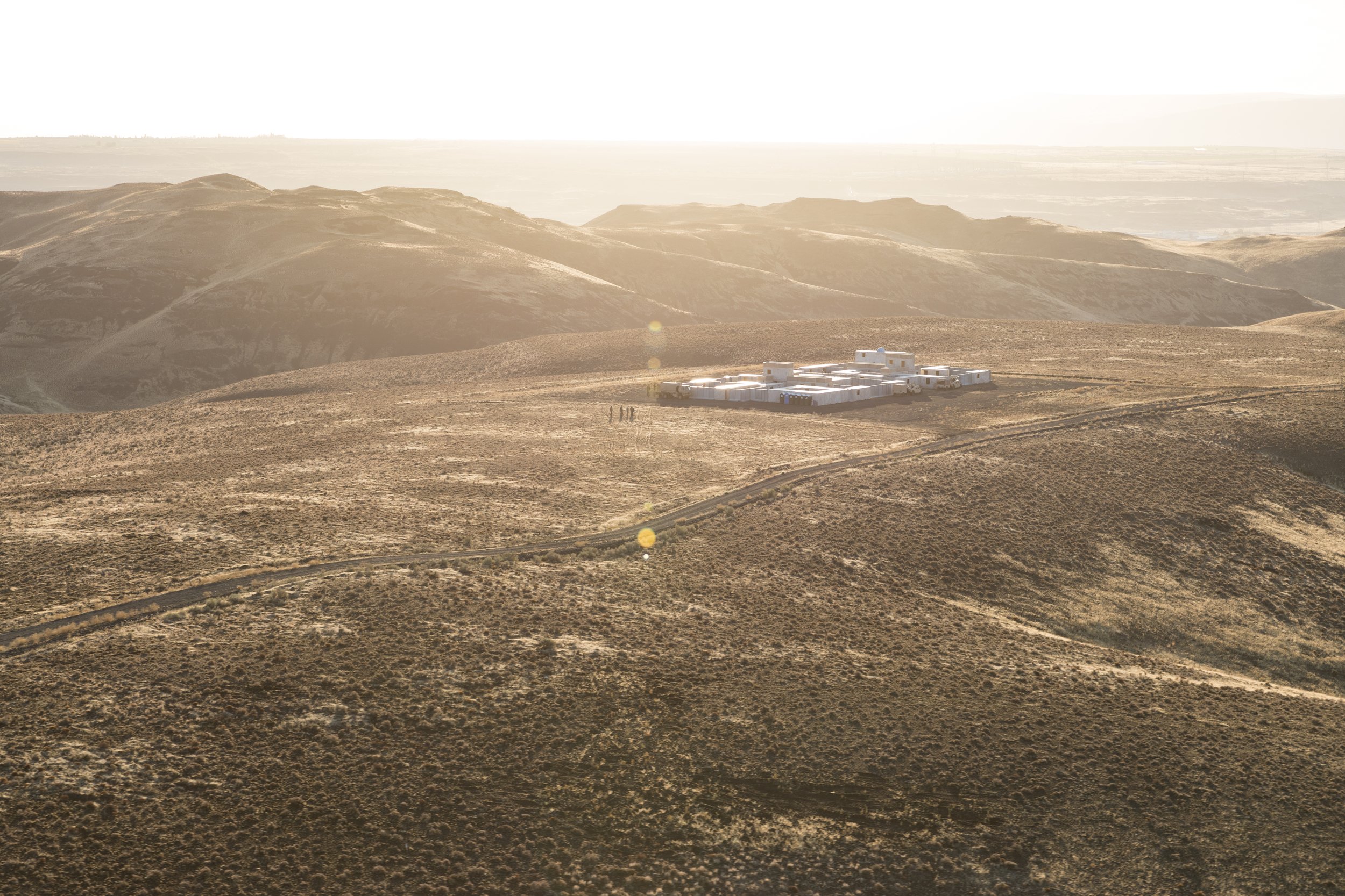 A small, white building complex located on a hilltop in a dry, hilly landscape with sparse vegetation, under a bright, hazy sky.
