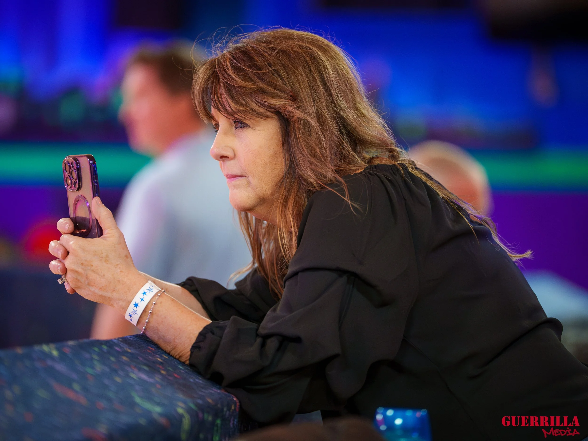 A woman with light brown hair, wearing a black top, is sitting at a table looking at her phone. The background is colorful and blurred, indicating an indoor setting. The woman has several bracelets on her wrist and is focused on her phone.