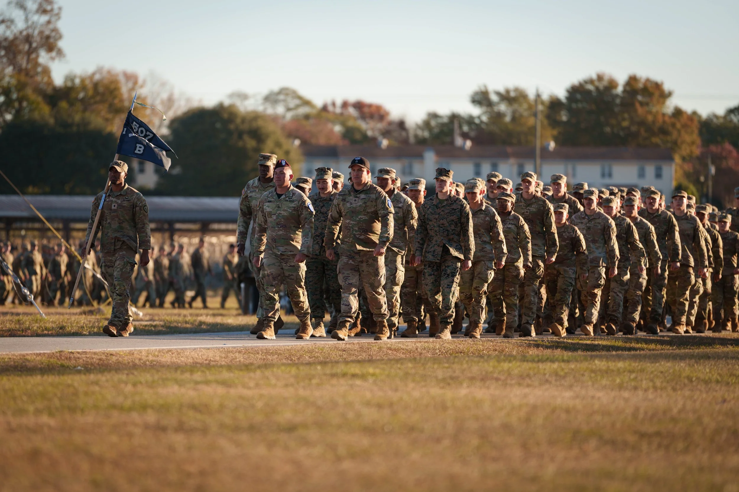 Military soldiers marching outdoors during daytime, wearing camouflage uniforms, with a flag bearer leading.
