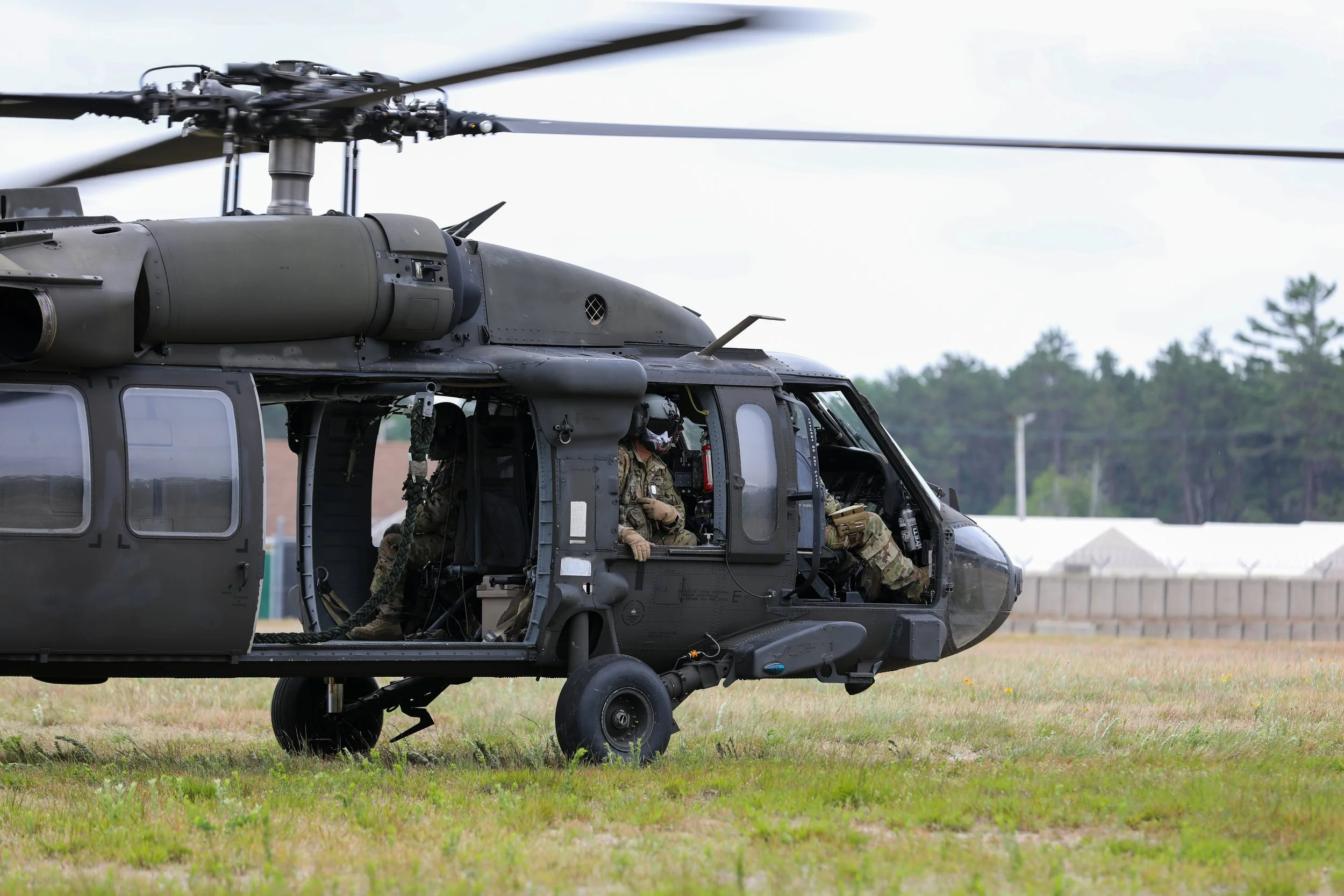 Military helicopter on the grass with soldiers inside wearing helmets and camouflage uniforms, with a background of trees and tents.