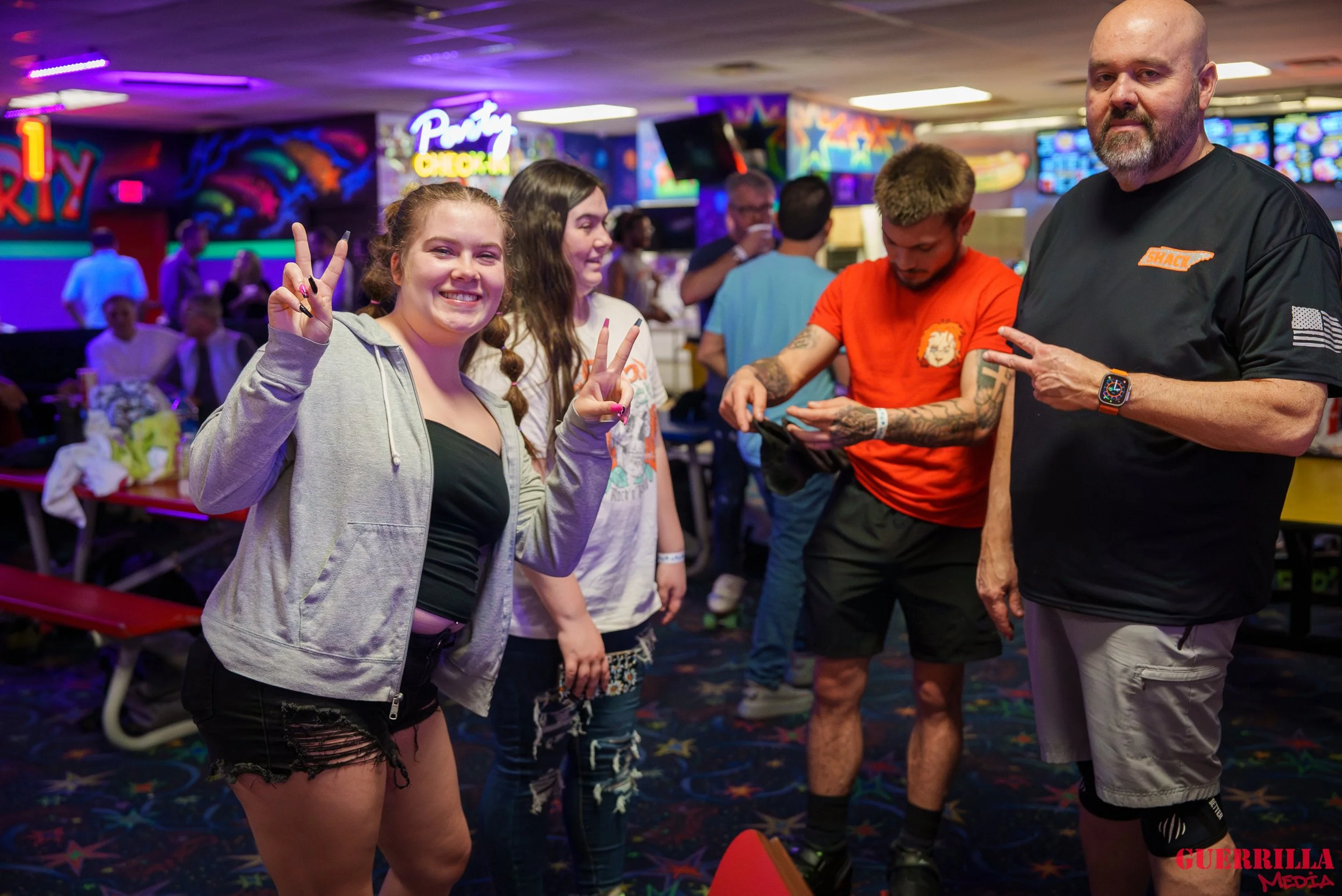 Group of young people at an indoor bowling alley, with neon signs in the background, some making peace signs, and others engaged with their phones.