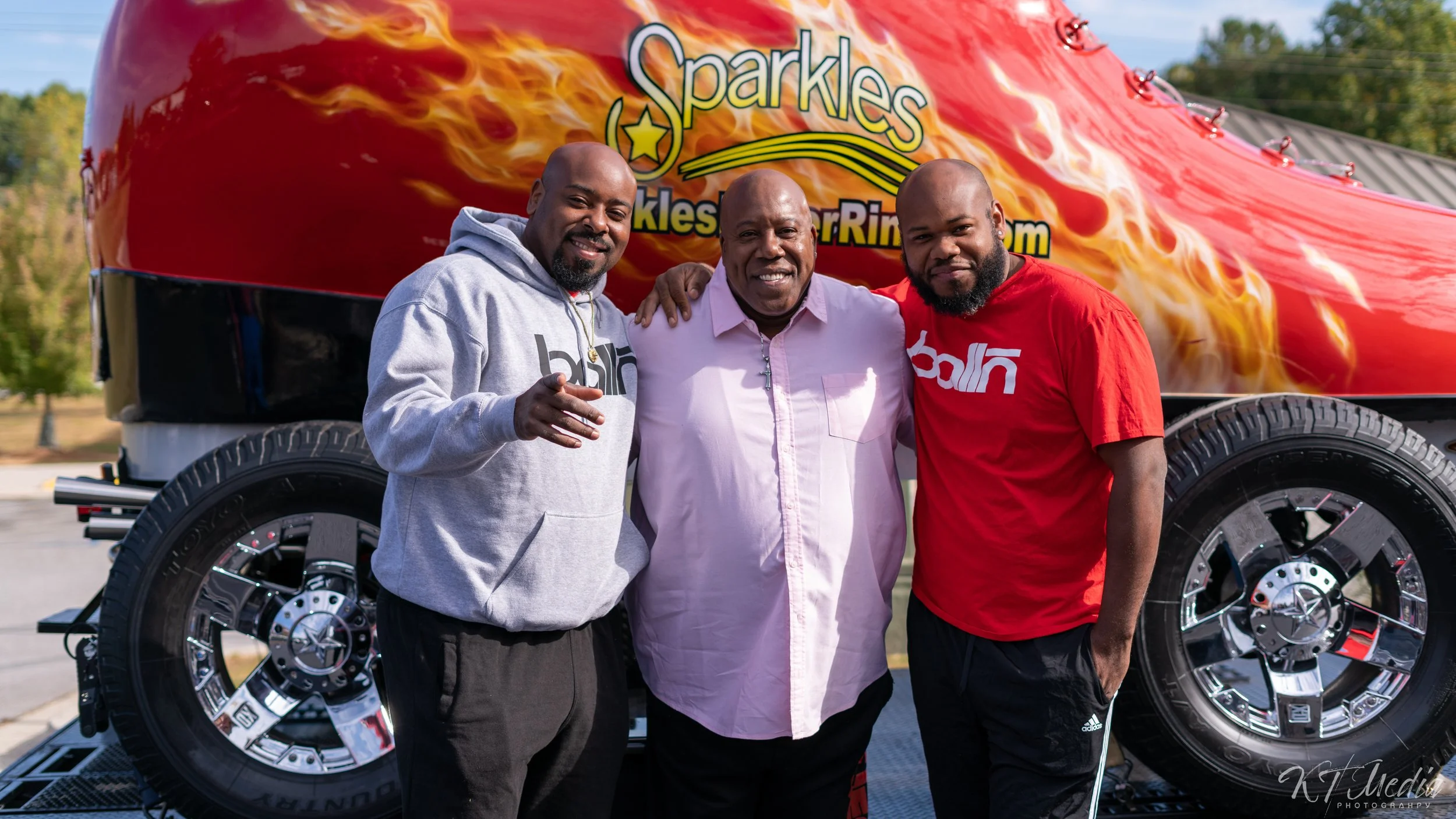 Three men standing close together outdoors in front of a large red and black monster truck with flames and the word 'Sparkles' on it. All are smiling, with arms around each other.