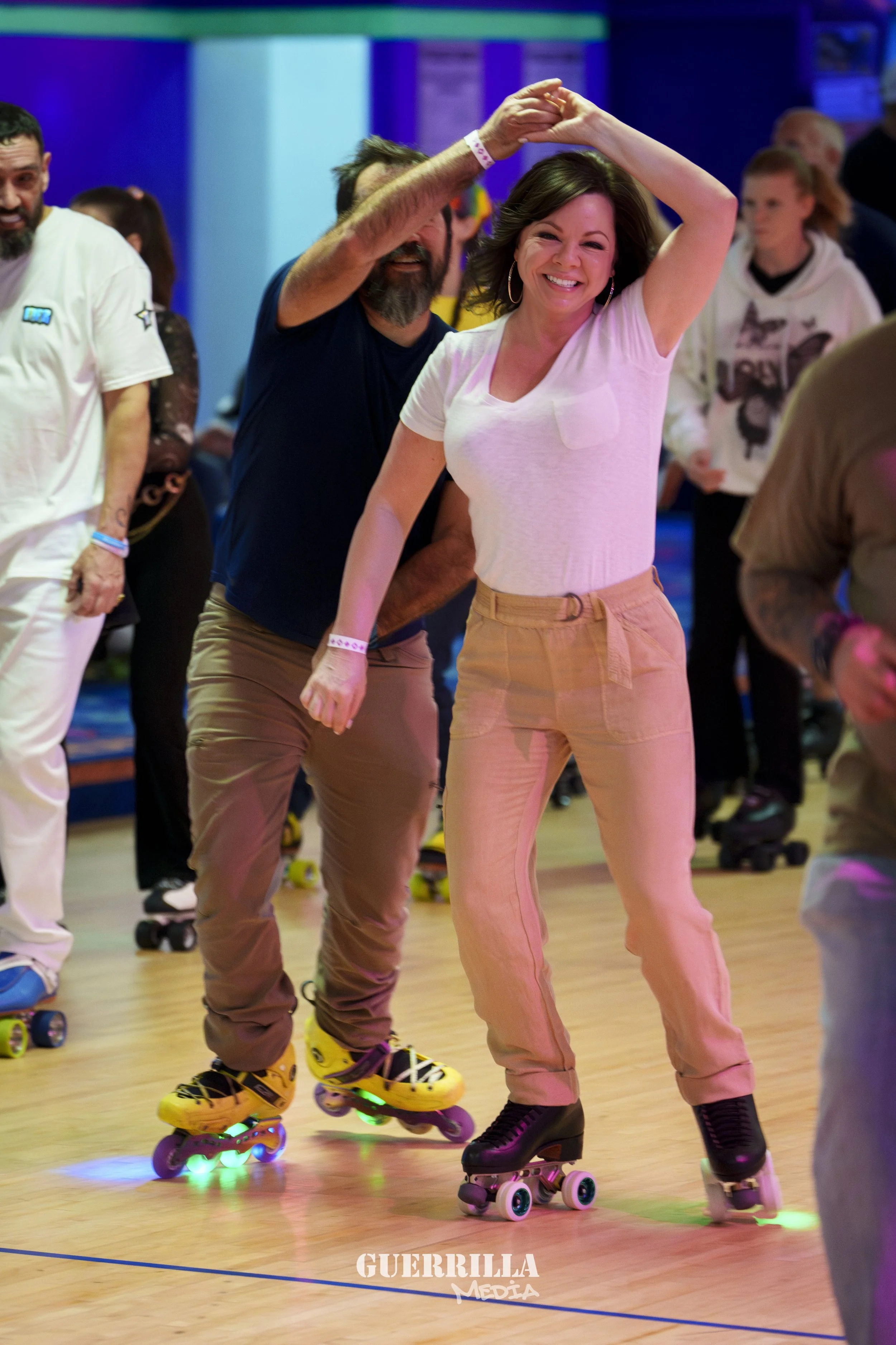 People roller skating at an indoor rink, with a woman smiling and dancing, wearing a light pink t-shirt and beige pants, skating alongside a man in a navy shirt and beige pants, wearing yellow roller skates with colored lights.