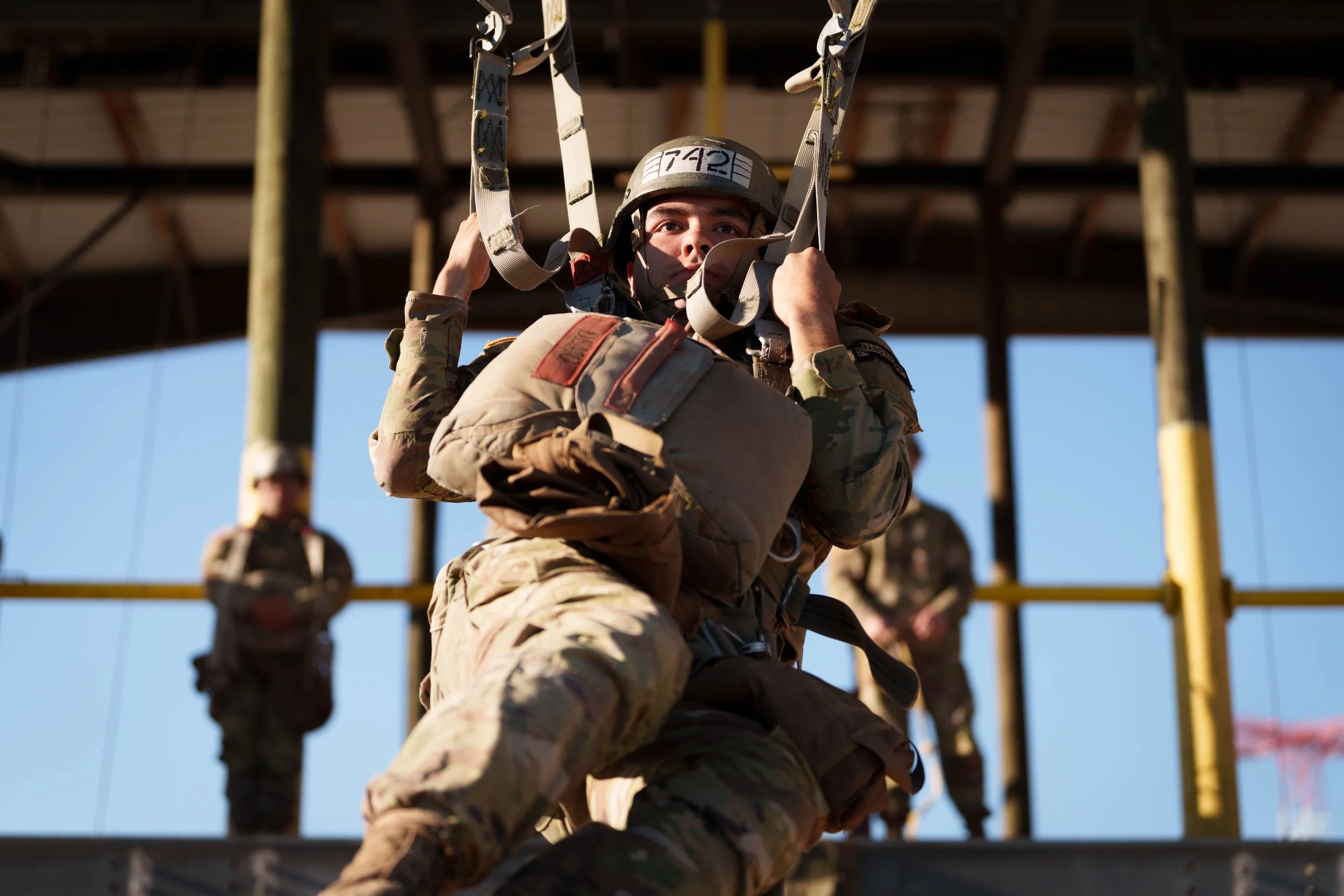 A soldier in camouflage uniform preparing to descend using a harness during a training exercise with several other soldiers in the background on a construction site.