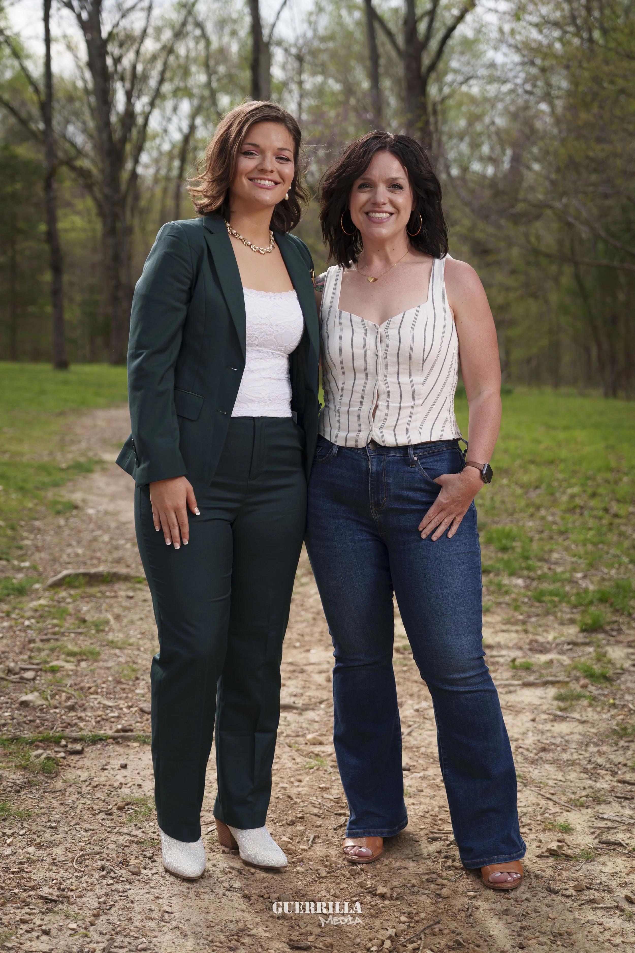 Two women standing on a dirt path in a wooded park, smiling at the camera.