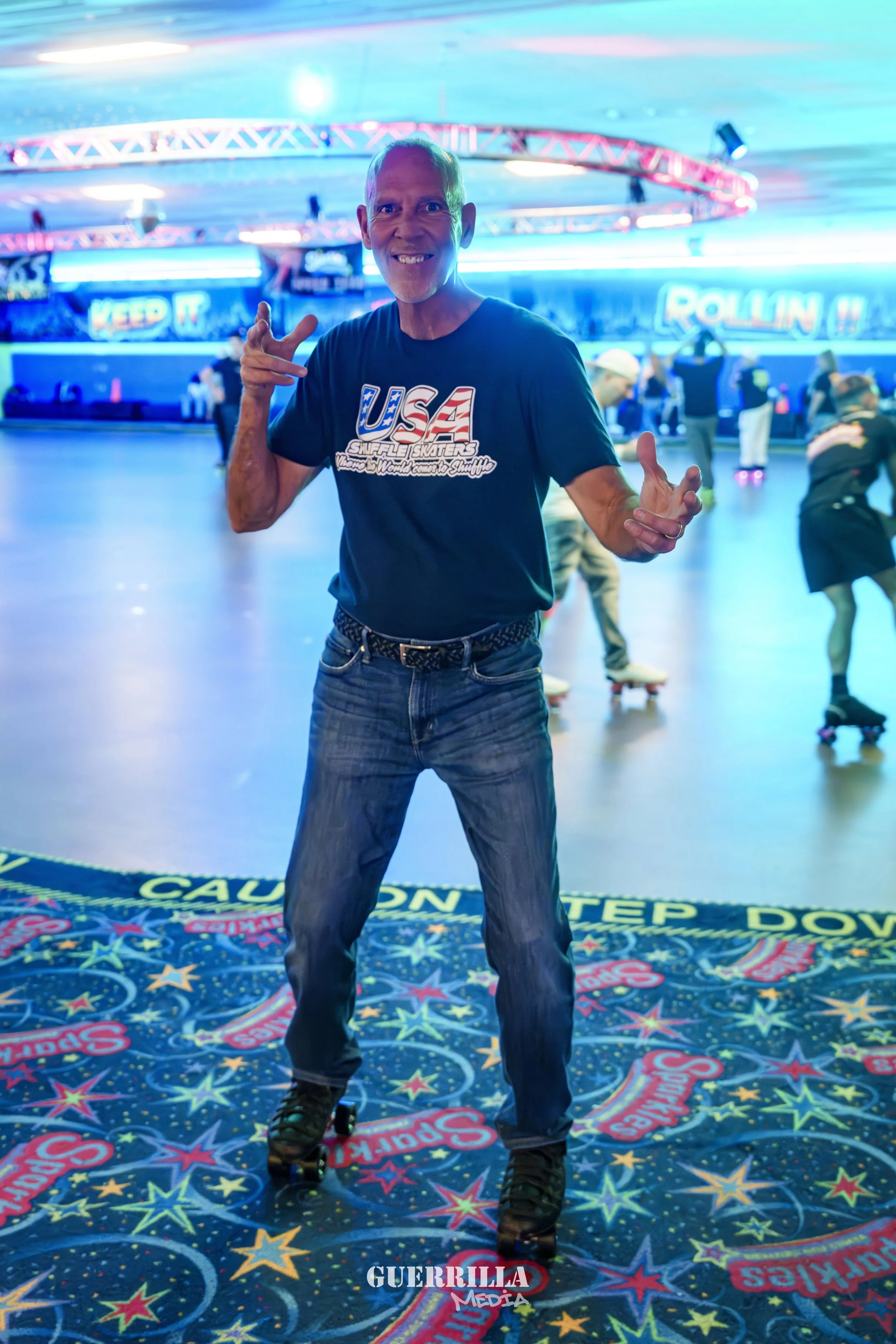 A man roller skating indoors at a roller rink with a neon and colorful starry floor mat, wearing a black t-shirt with USA and skateboard graphics, and surrounded by other skaters in the background.