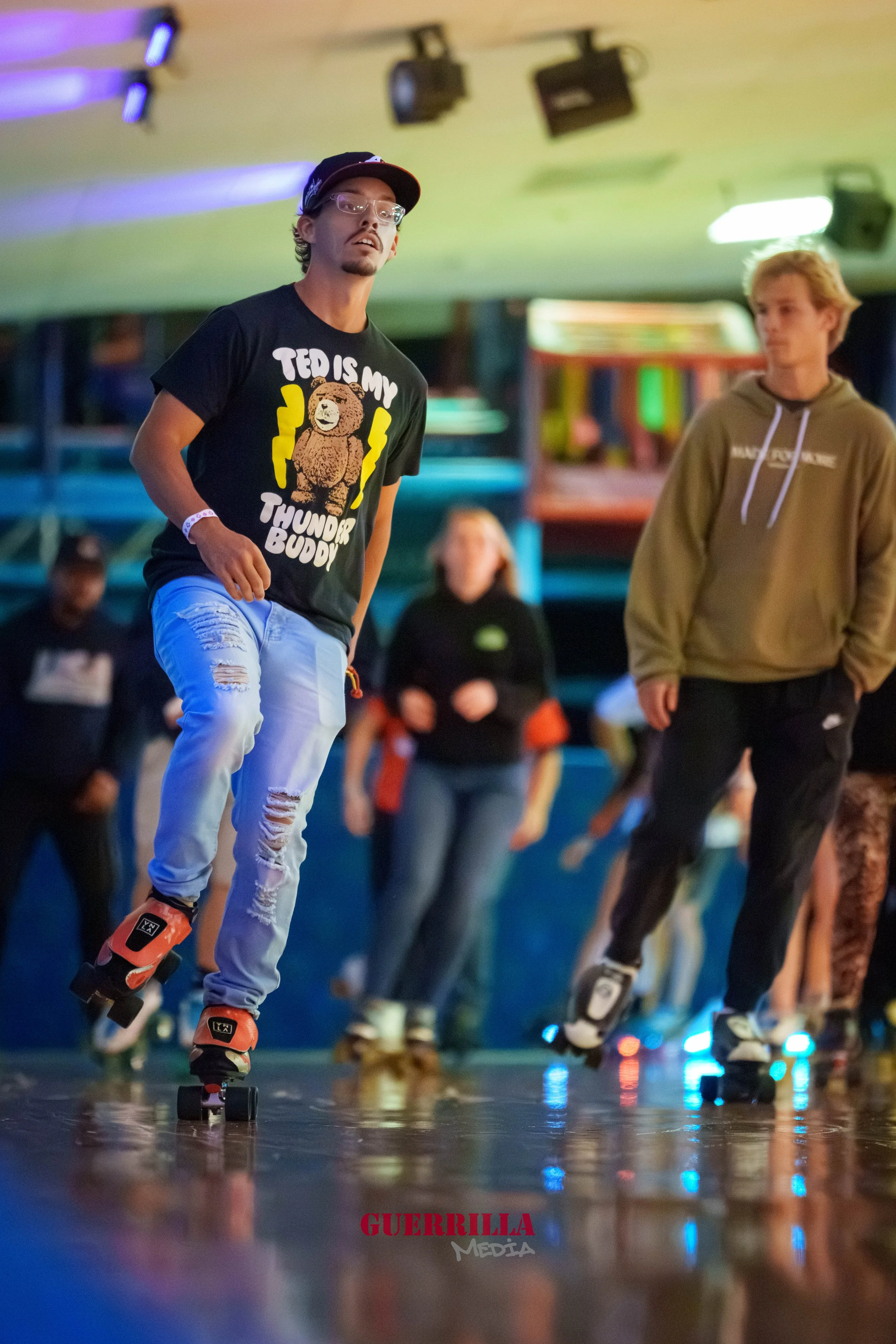 Young man roller skating in an indoor skating rink, wearing a black T-shirt with a bear graphic and the text 'Ted is my thunder buddy,' and light ripped jeans, with other people in the background also skating.