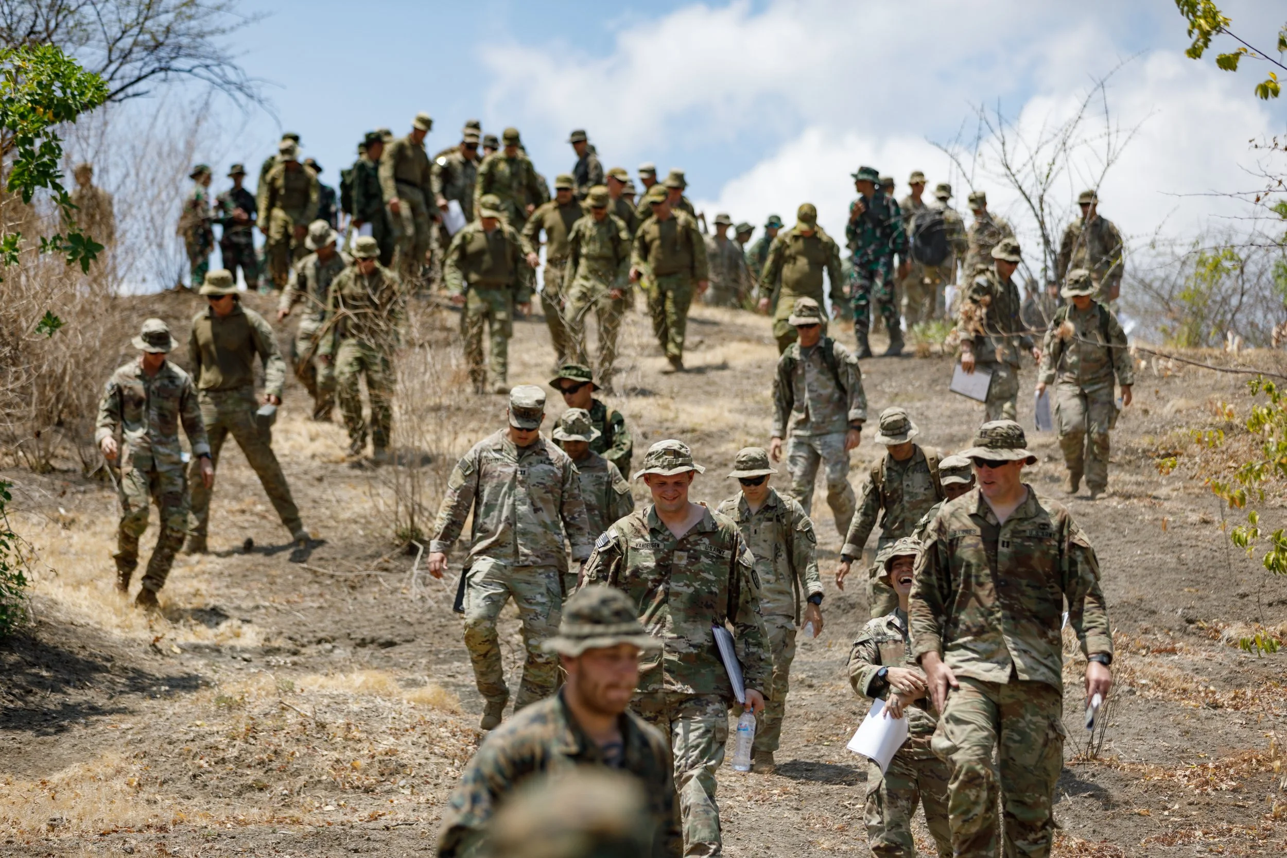 A group of soldiers in camouflage uniforms and hats walking down a dirt path, some carrying documents and water bottles, in a dry outdoor setting with sparse vegetation.