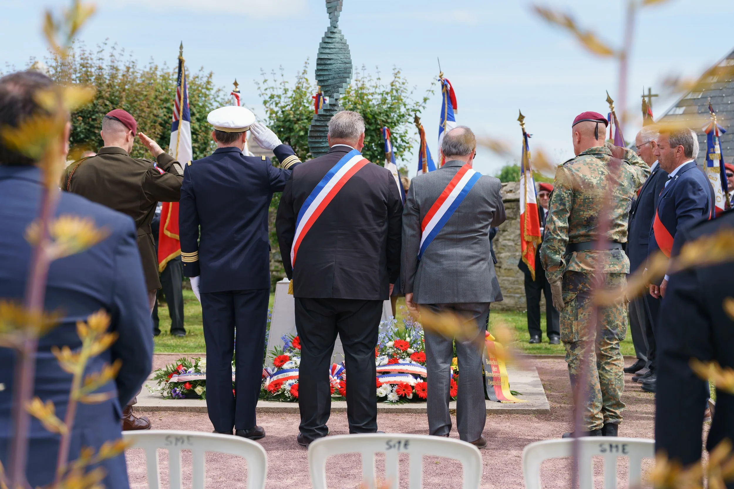 A group of people, including military personnel and officials, are standing with their backs to the camera during a memorial ceremony. They are paying respects at a monument adorned with flowers and wreaths. Several flags are displayed behind them, a