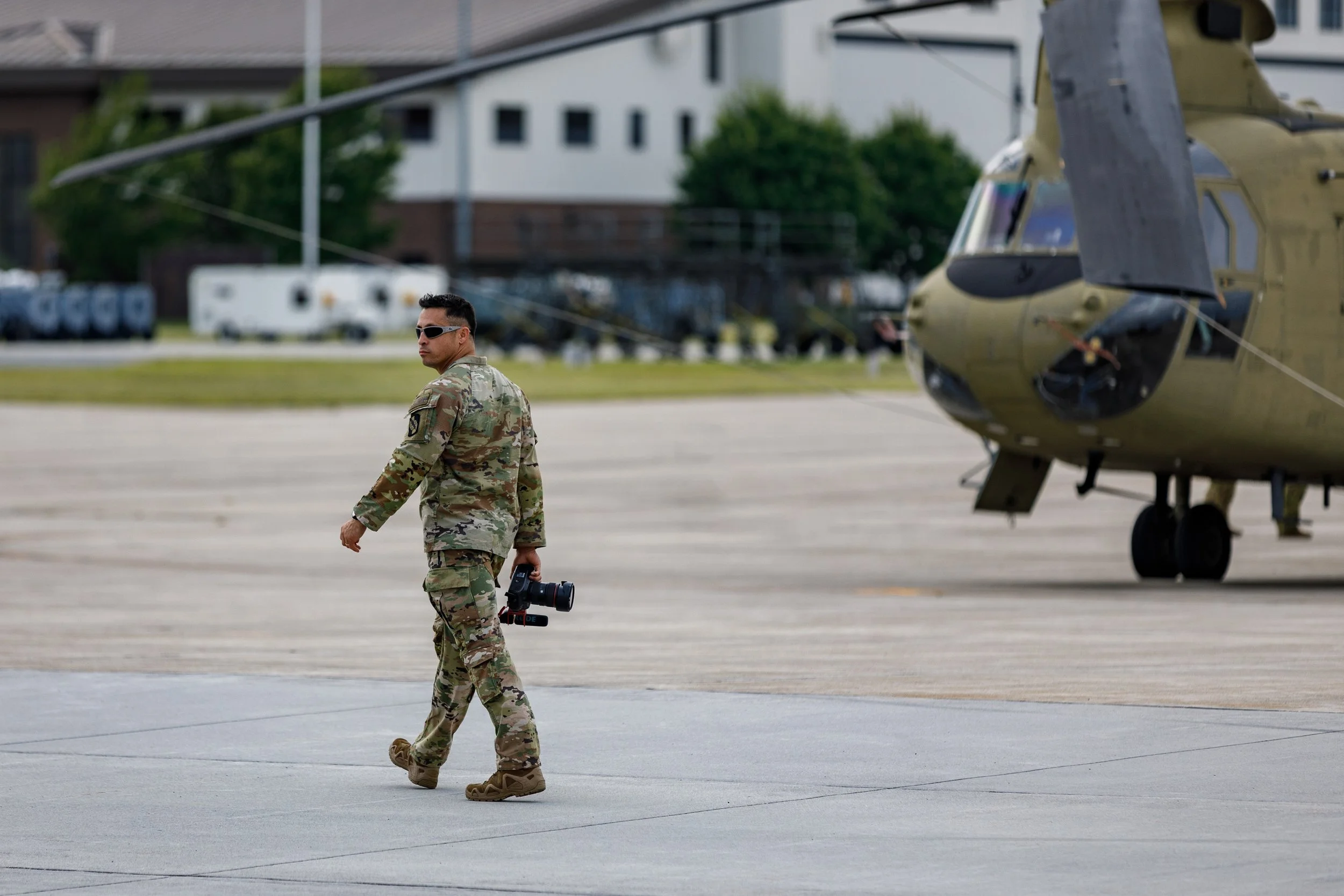 A soldier in camouflage uniform and sunglasses walking on an airstrip holding a camera, with a helicopter in the background.