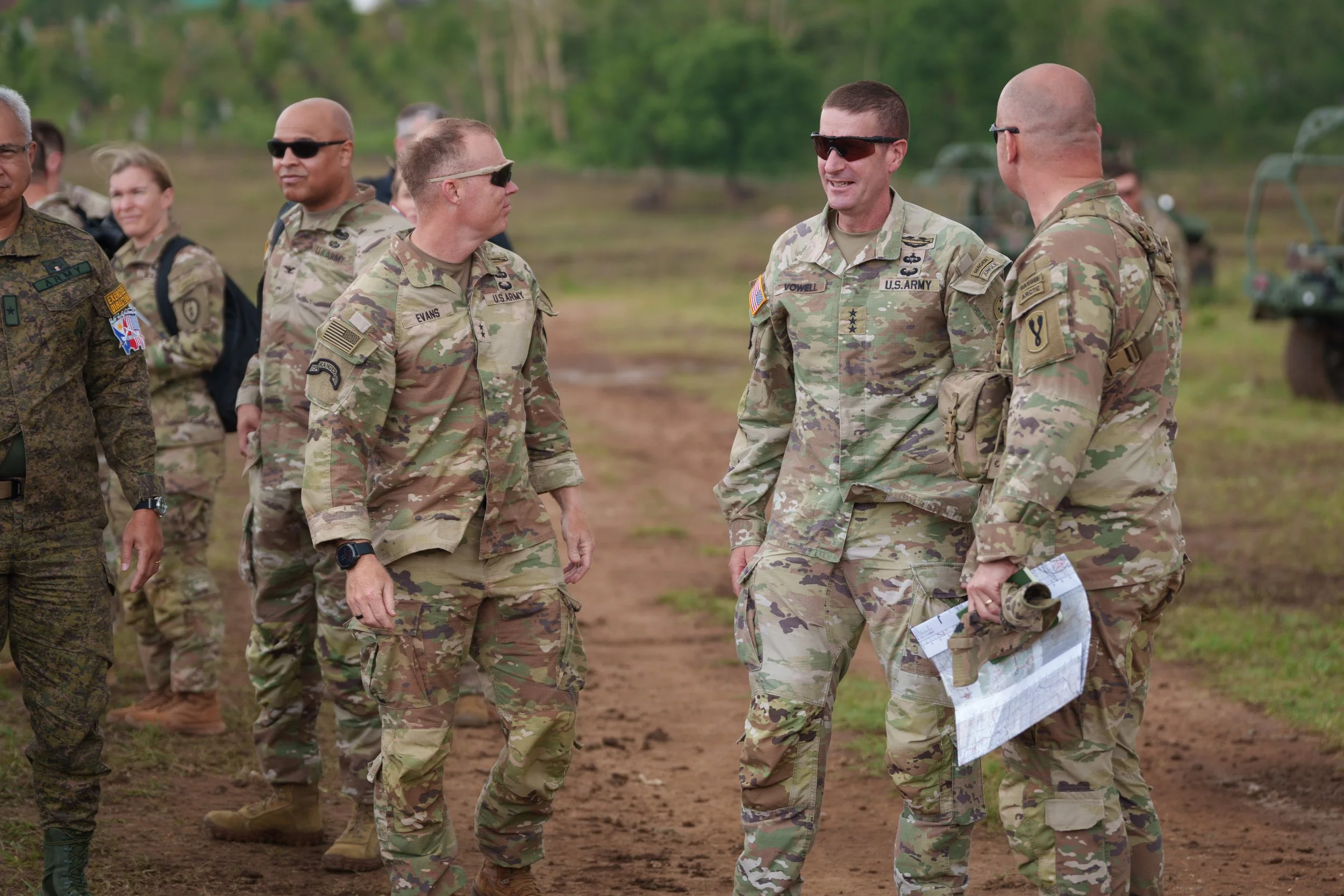 Group of U.S. military personnel in camouflage uniforms talking outdoors on a dirt path, with trees and military vehicles in the background.
