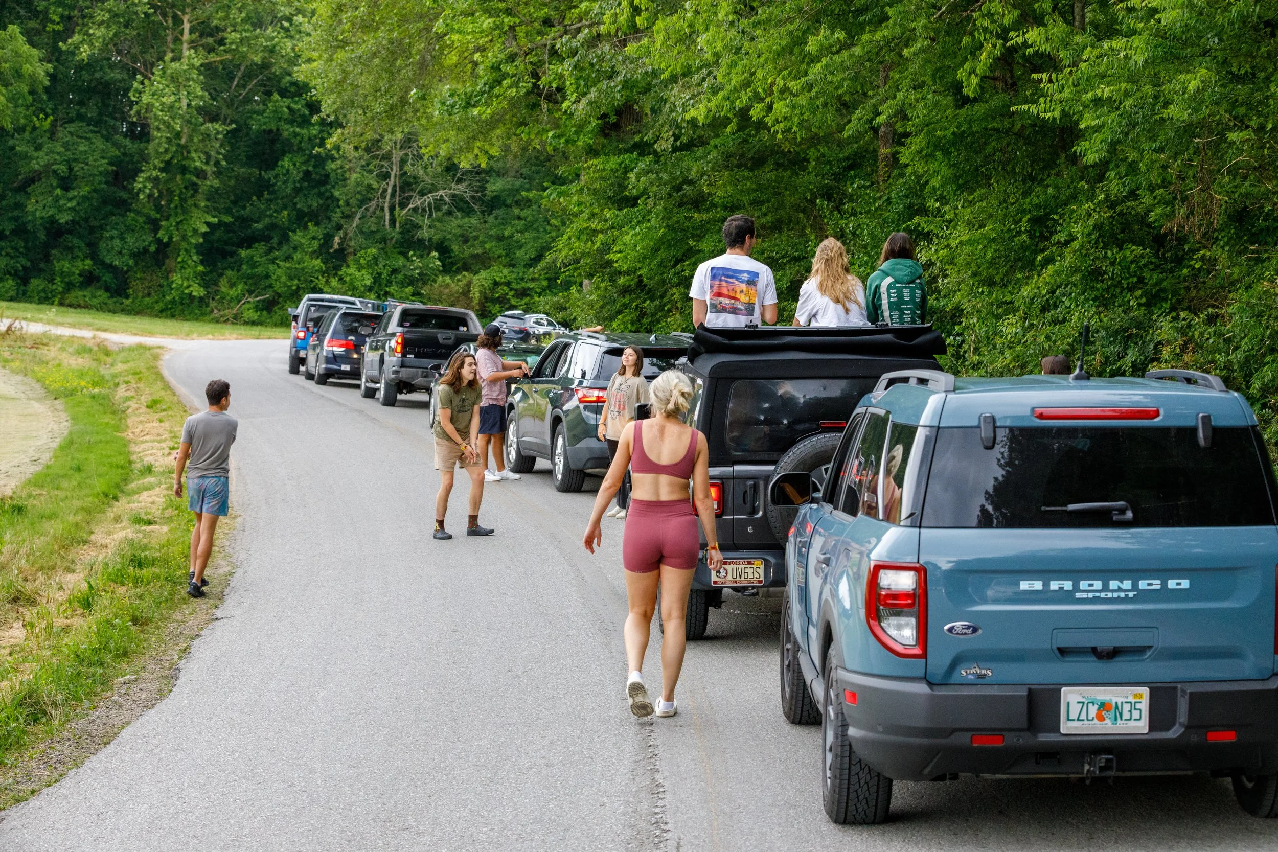 People standing and sitting on cars parked along a rural, tree-lined roadside, with some engaging in conversation and others looking at the surroundings.