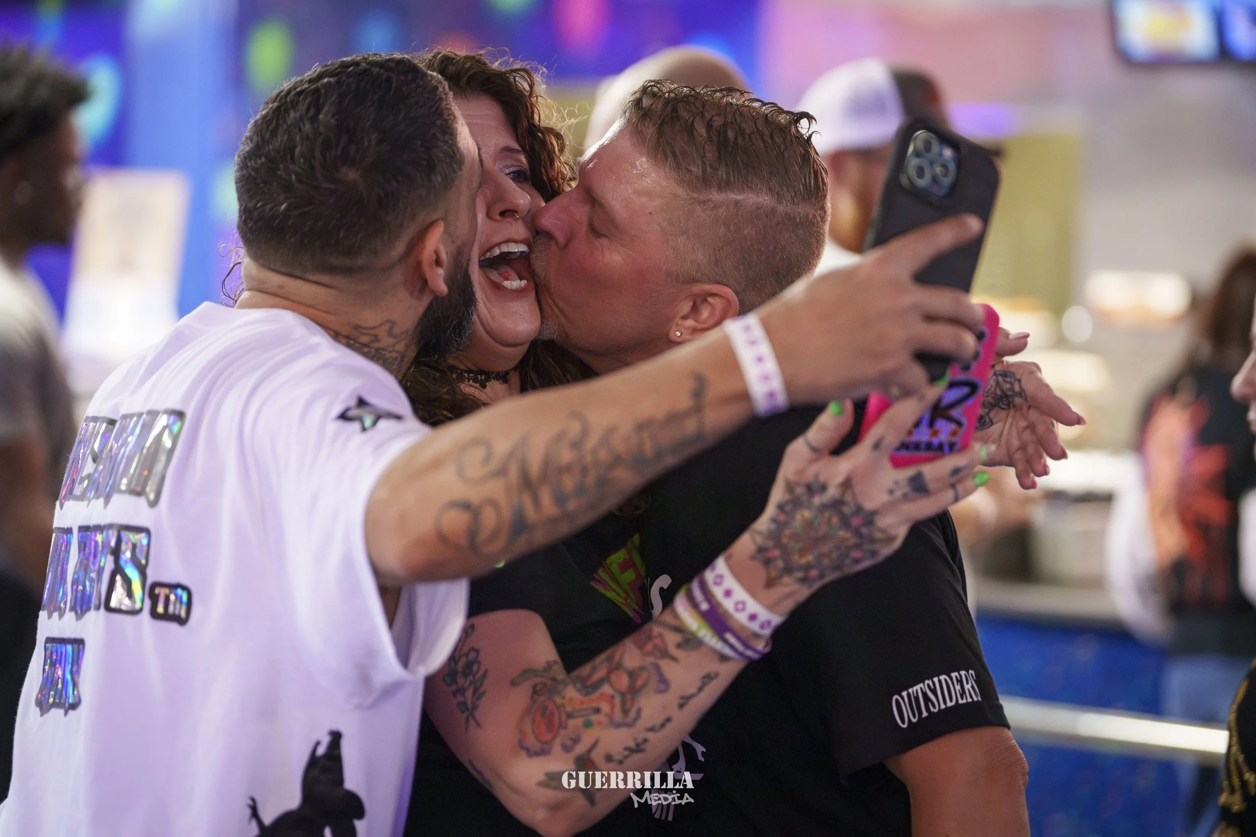 Three people taking a selfie together at an indoor event, with a woman in the middle being kissed on the cheek by a woman on the right and a man on the left holding the phone. They are smiling and appear happy.