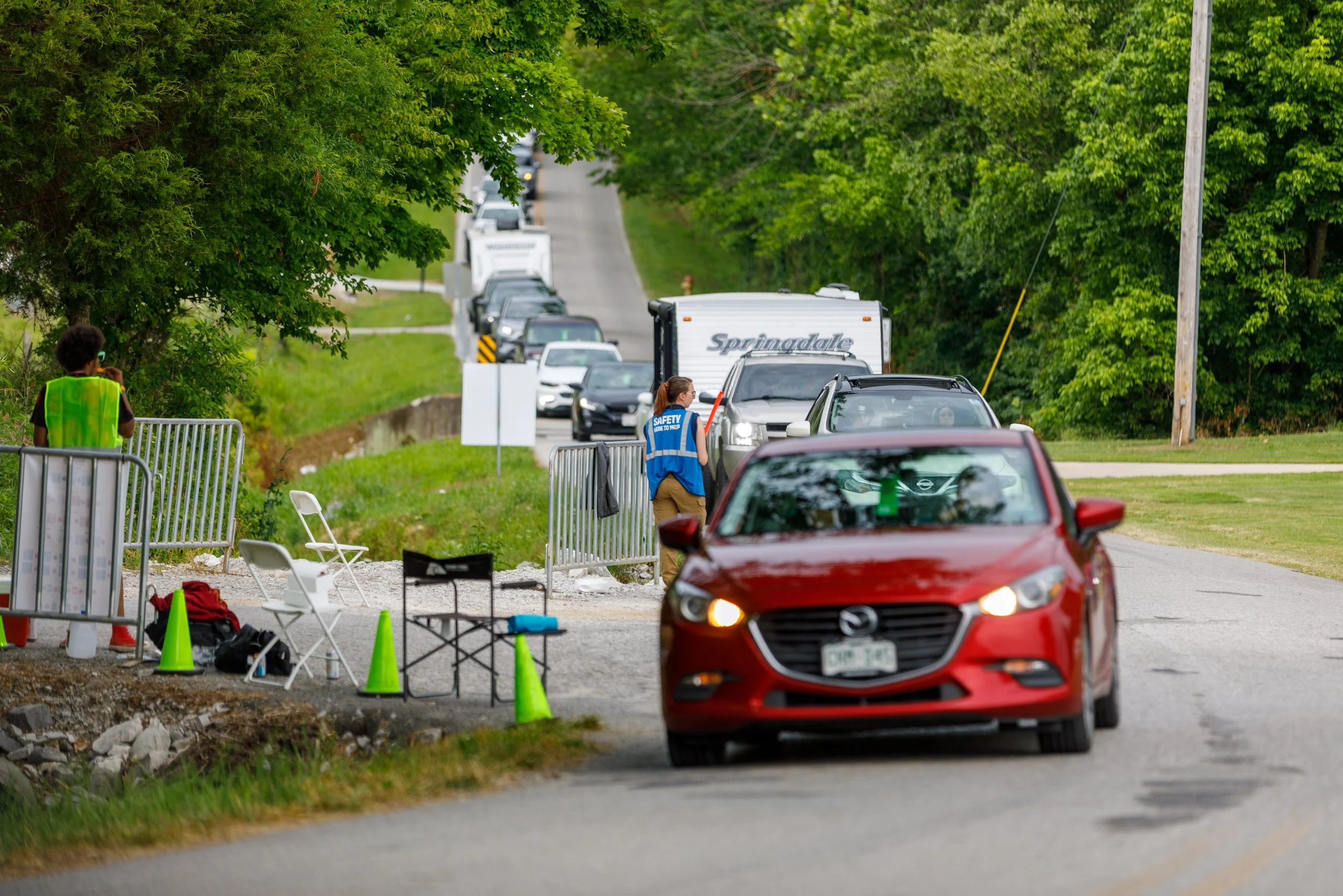 Line of cars on a rural road waiting at a checkpoint with safety personnel and barriers.