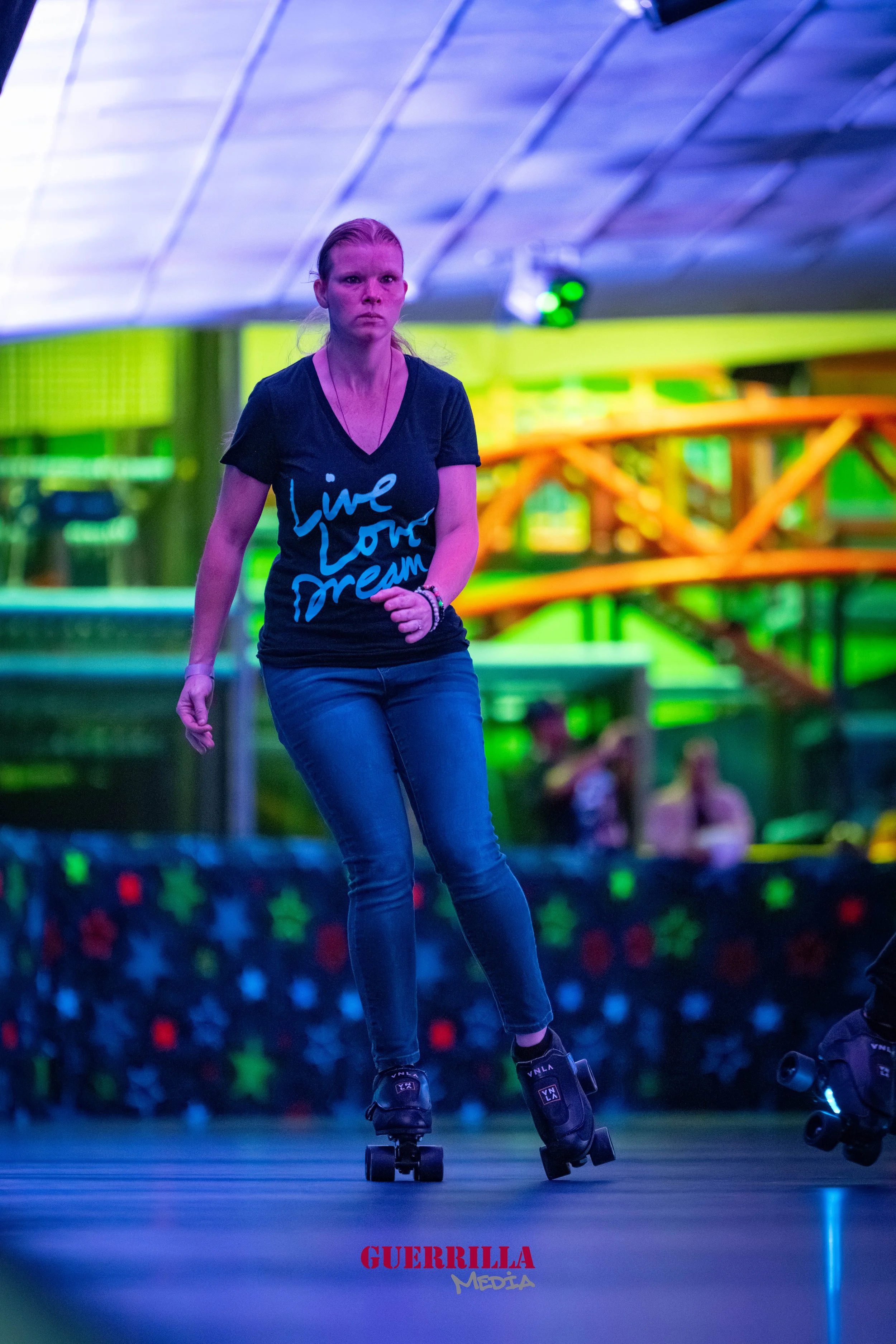 A woman roller skating indoors under colorful neon lights, wearing a black t-shirt with the words 'Live Love Dream' written on it and dark jeans.