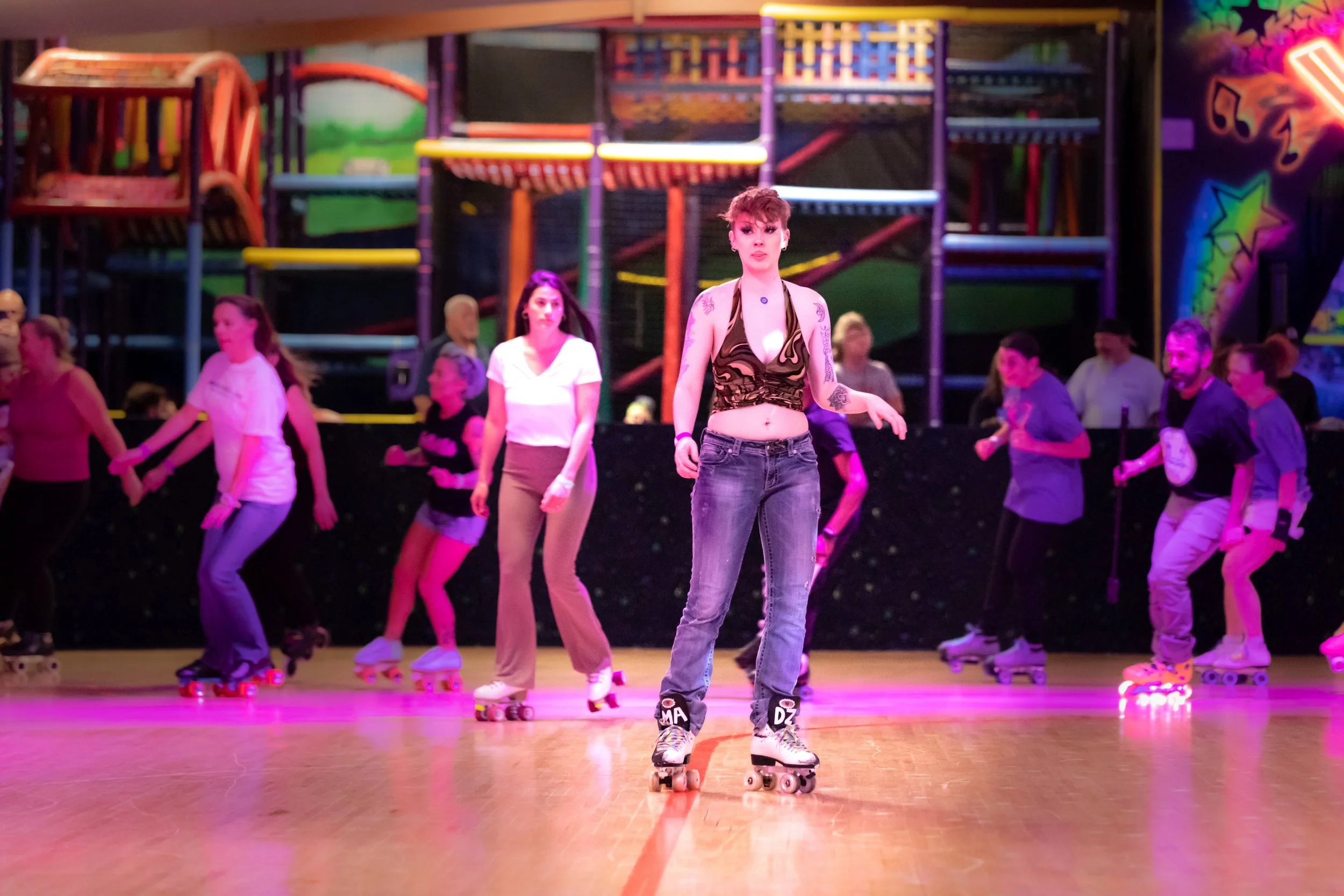 A group of people roller skating in an indoor roller rink with colorful neon lights and a funhouse in the background.