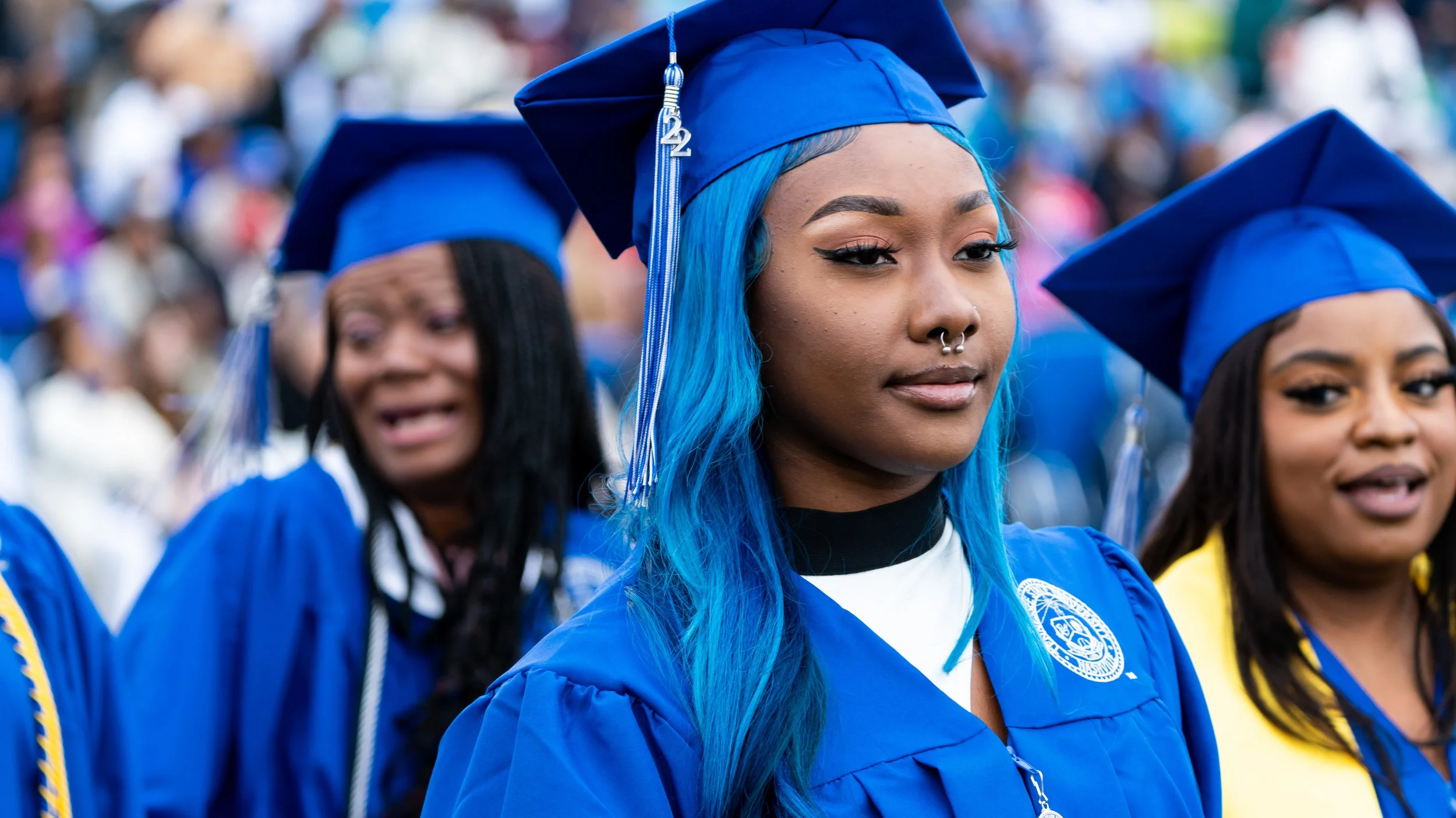Group of young women in blue graduation caps and gowns at a graduation ceremony, with one woman in focus having long blue hair, nose piercing, and a confident expression.