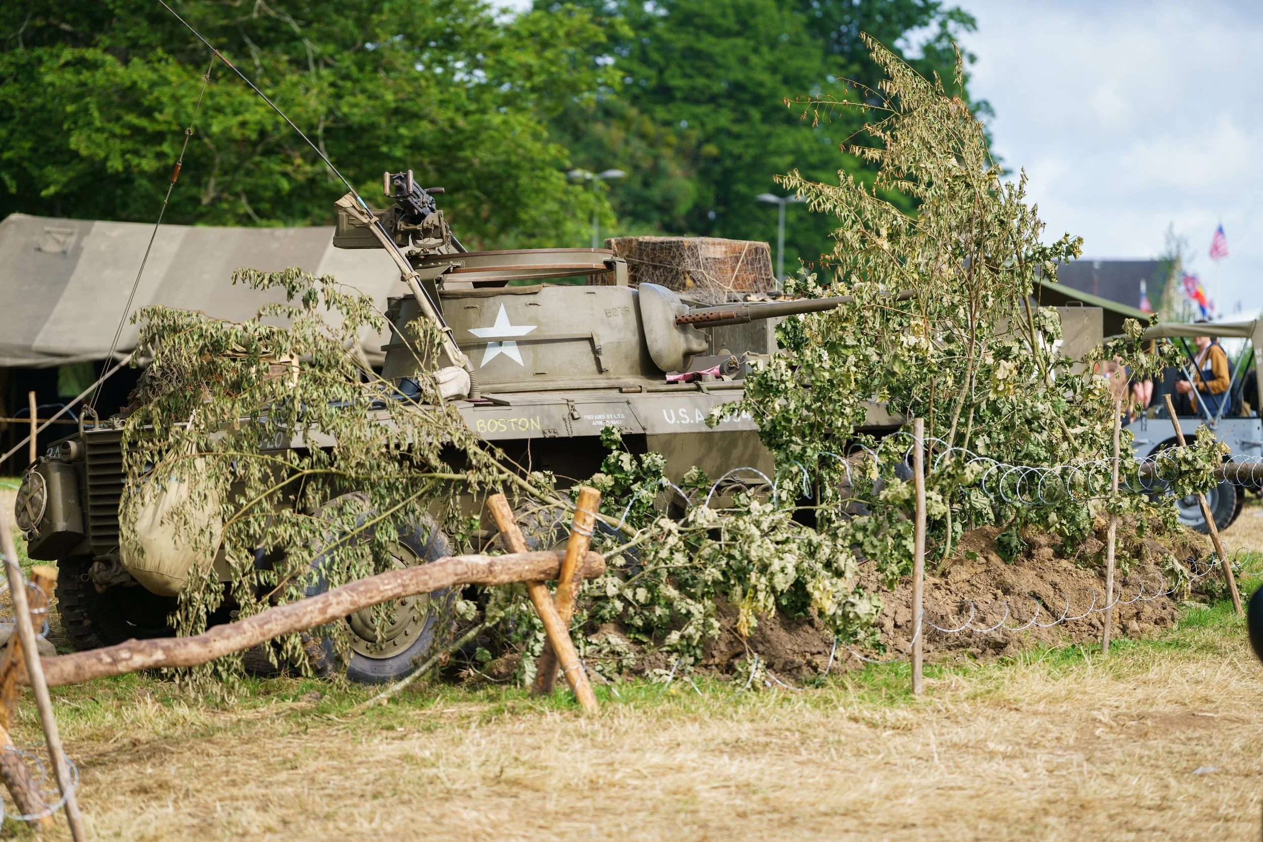A military tank is behind a barbed wire fence with branches and leaves on or near it, at an outdoor event or display. There are tents and people in the background.