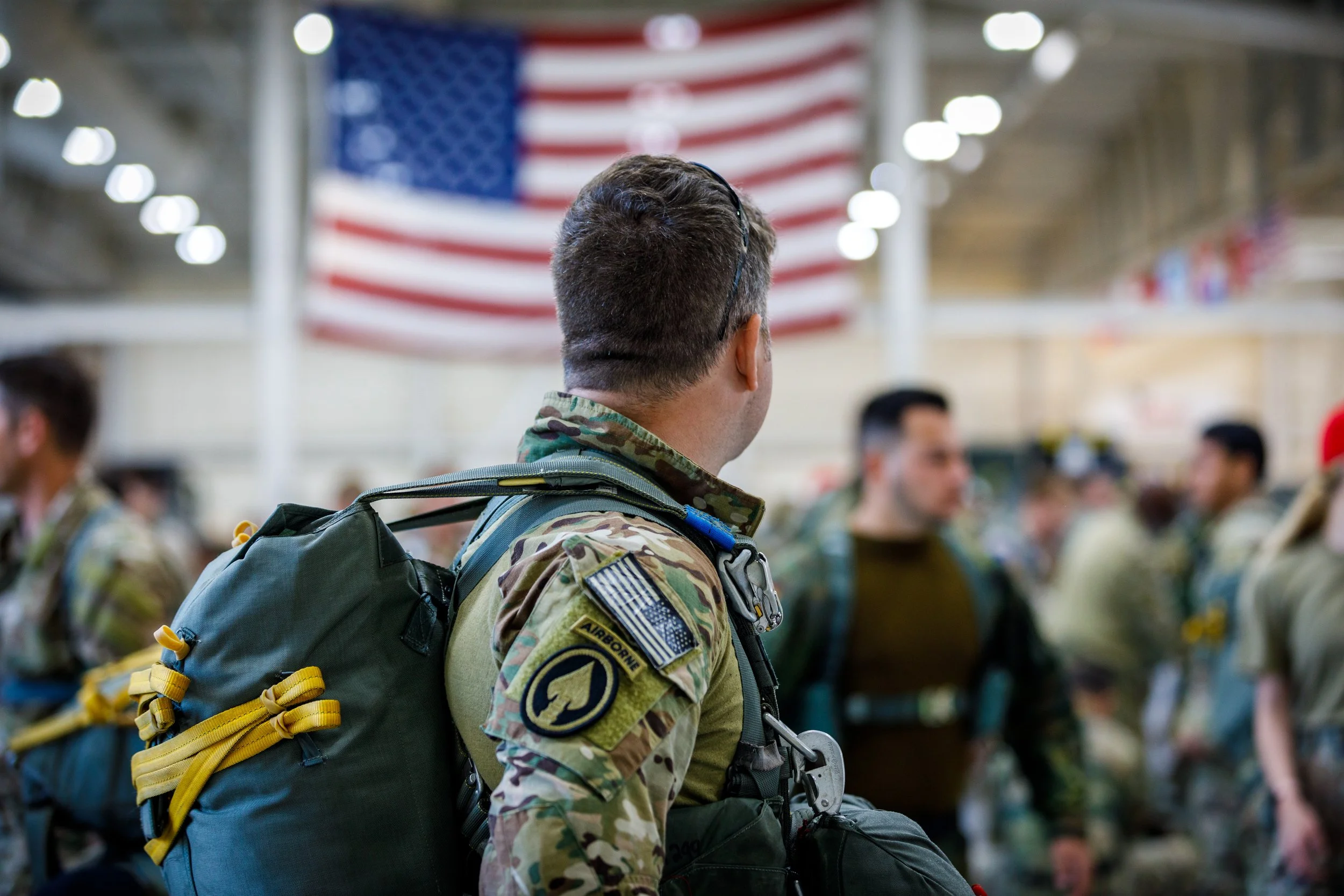 A soldier in camouflage uniform standing in a crowded indoor space with an American flag hanging in the background.